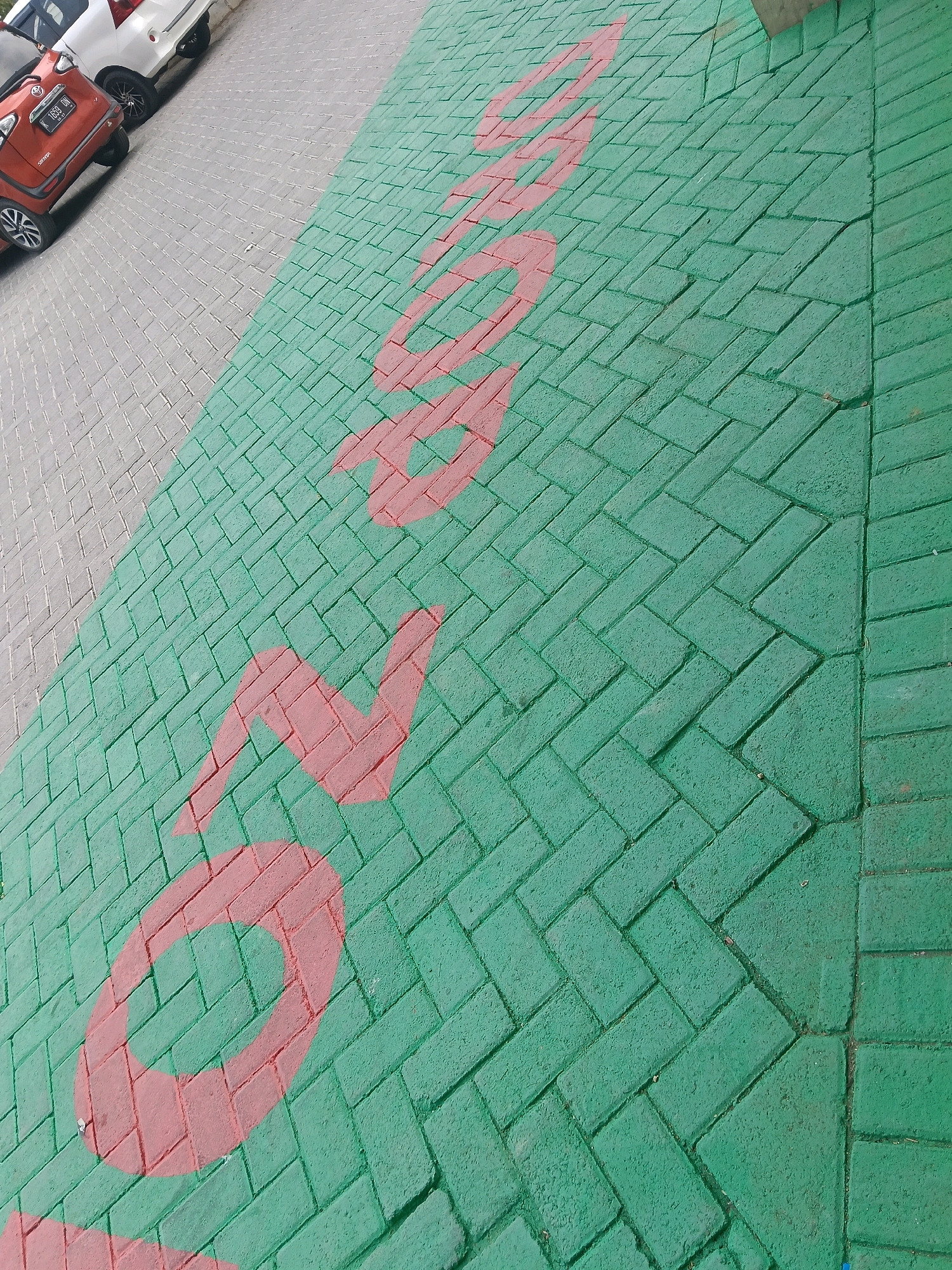 The image depicts an outdoor paved area with interlocking brick pavers. The pavers are predominantly painted a vibrant green, with some sections appearing weathered. Overlaid on the green pavers are large, stylized red painted characters, forming what appears to be text or a logo. The characters are broad and rounded, with a bold outline. To the left of the green paving, there is an area of grey pavers. Partially visible on the grey pavers are the rear ends of two cars. One car is orange and has a license plate that reads "K 1659 DN". The other car is white. The scene appears to be in a public or commercial area, possibly a parking lot or walkway, in Balun, Indonesia.