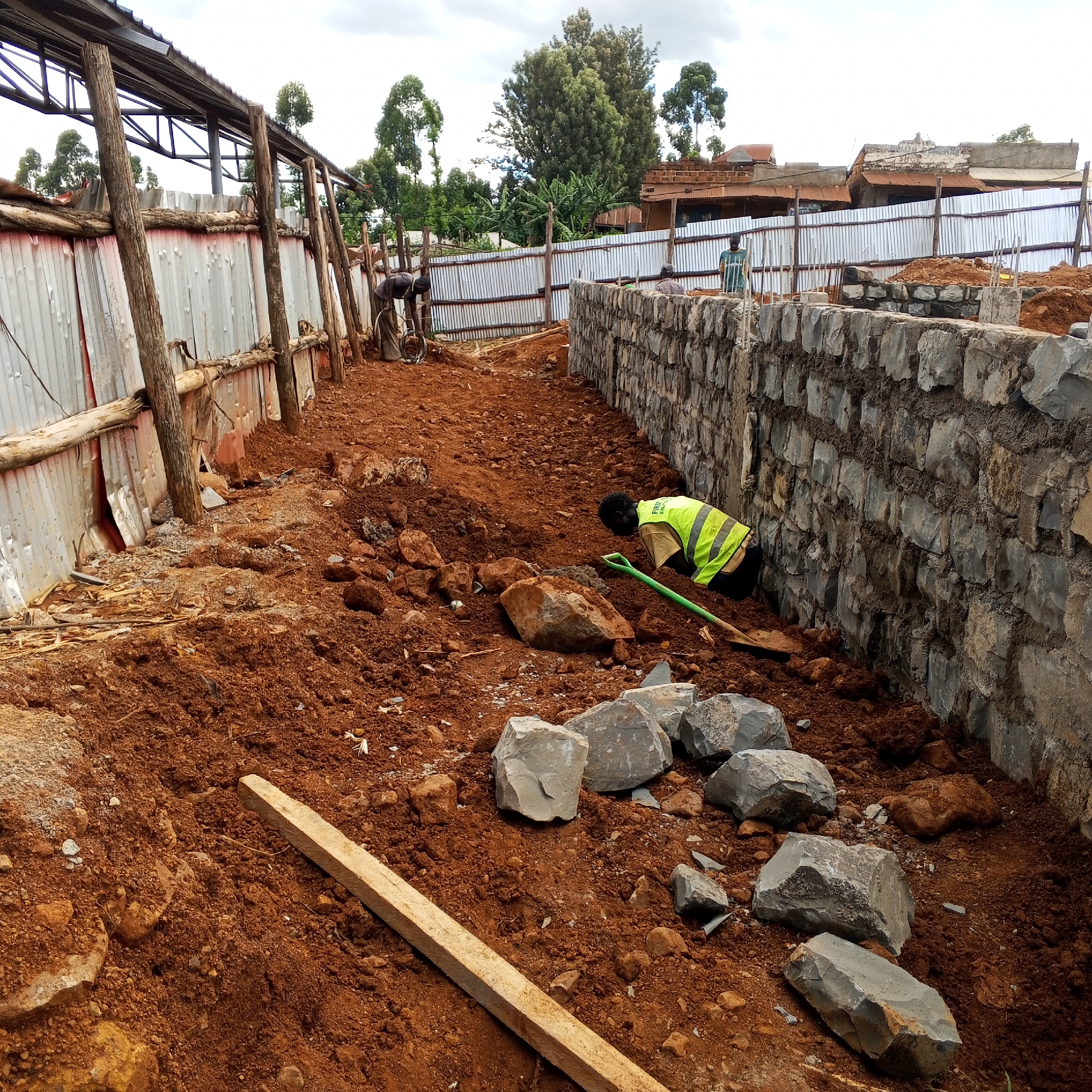 This image captures an active construction site in Borabu Chitago ward, Kenya, under an overcast sky during daytime. The foreground is dominated by reddish-brown earth scattered with numerous large, rough-hewn stones and smaller rocks.
Three individuals are visible, all engaged in work. In the immediate foreground right, a worker wearing a yellow-green high-visibility vest (with partially visible text "FIBER SOLUTI") over a light shirt and dark trousers is bent over, holding a green-handled shovel, appearing to be digging or moving earth and stones. To their right, a partially completed wall made of rough-hewn stones is visible, with mortar visible between the stones.
On the left side of the frame, a structure with a metal roof frame and corrugated metal sheet walls is being erected or reinforced. Another worker in dark clothing is bent over near this structure, working close to a wheelbarrow or similar equipment. Further in the background, a third person, wearing a patterned shirt, stands near the ongoing stone wall construction.
The background reveals a mix of lush green vegetation, including tall, slender trees and bushier plants, typical of the region. Beyond these, several residential buildings with corrugated iron roofs and brick walls are discernible. On one distant, faded building, faint text "WOMEN SELF-HELP SHOP" can be seen. The overall scene depicts ongoing manual labor and construction activities in a rural or semi-urban setting.