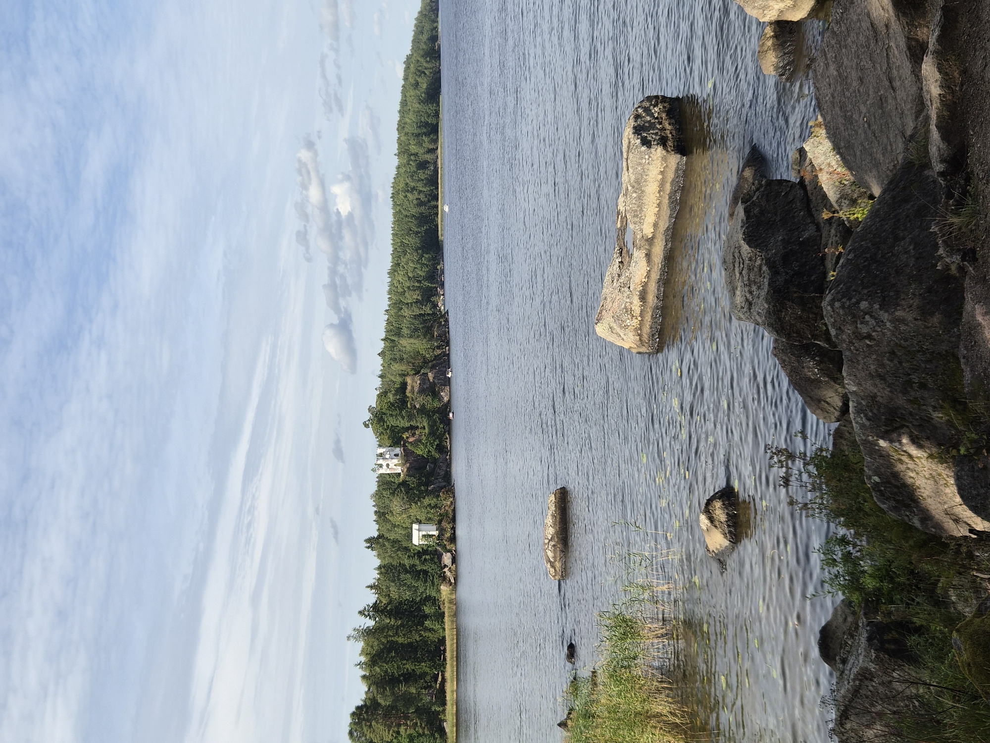 The image depicts a serene waterscape with a rocky shoreline in the foreground. A calm body of water stretches towards a densely forested island in the background. On the island, amidst the trees, two white structures are visible: a smaller, square building and a larger, multi-story building situated on a higher elevation. Several boats are anchored or moving in the water near the island. The sky above is partly cloudy, with a mix of white and gray clouds against a pale blue background. The overall scene suggests a tranquil, natural environment with some man-made structures integrated into the landscape. The Vyborg Bay in Vyborg, Russia, is a likely location for such a scene, characterized by its islands, forests, and historical buildings.