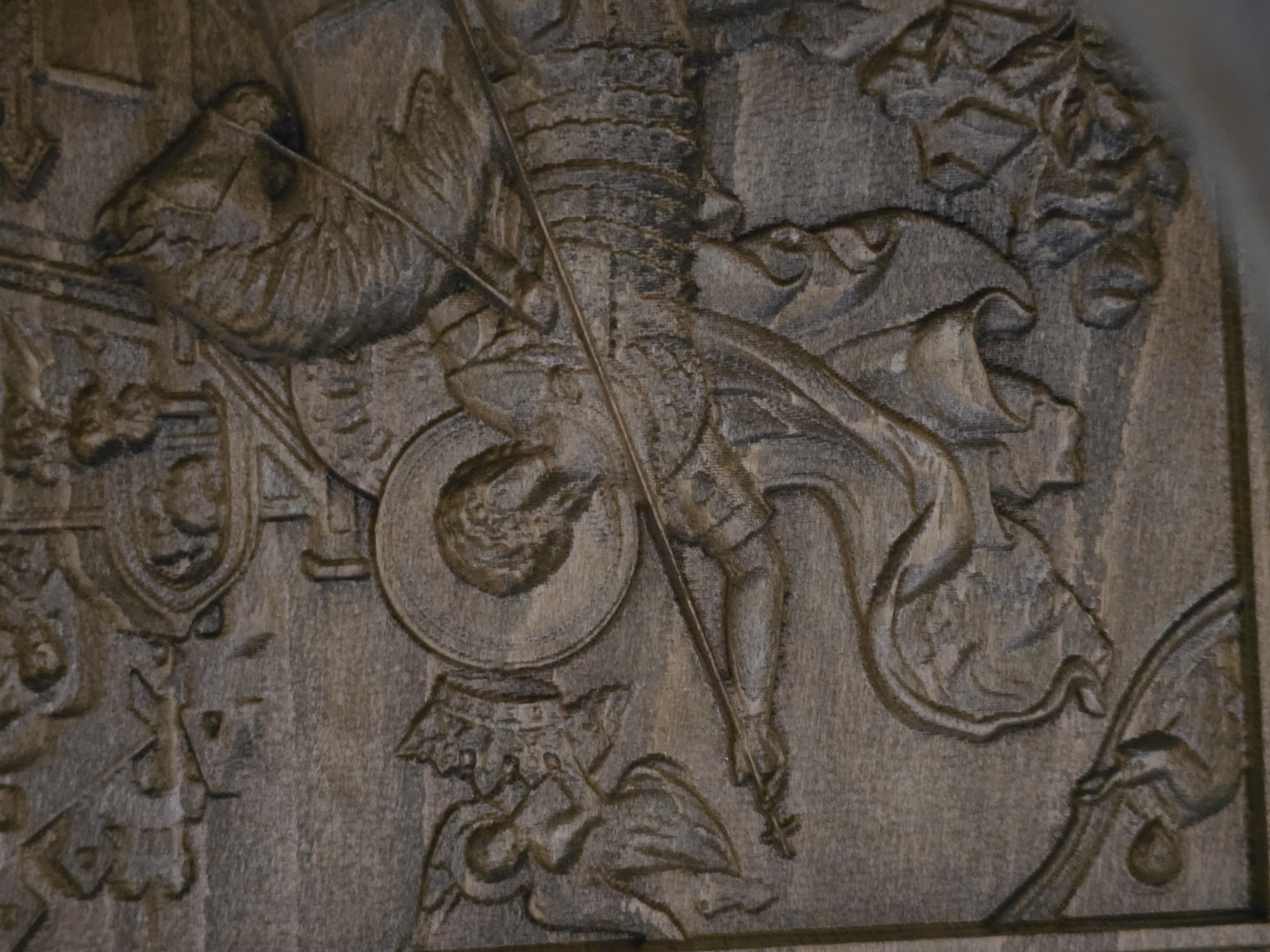 This media file displays a detailed relief carving, possibly made of wood or stone, characterized by its textured surface and monochromatic brown and grey tones. The central subject is a dynamic figure, likely a warrior or knight, depicted in elaborate armor. He is shown holding a long staff or lance diagonally across the frame, with what appears to be a small cross or finial at its tip.
The scene features multiple intricate elements:
* In the upper left, a large winged creature, possibly a griffin or eagle, is carved with visible feathers and a beak.
* Below the central figure, towards the bottom left, a smaller winged figure, resembling a putto or cherub, is shown wearing a distinct coronet or crown-like headpiece. This figure appears to be beneath the main warrior.
* To the left of the main figure, several heraldic shields or coats of arms are visible, adding to the historical or allegorical nature of the carving.
* On the right side of the composition, flowing draperies or fabric are carved with depth, and in the upper right corner, intricate foliage with leaves and berries can be seen.
The carving exhibits significant depth and detail in the armor, wings, and decorative elements. There is no visible text. The overall impression is that of a complex, aged artistic piece, possibly depicting a mythological, religious, or historical event, rendered in a classical or baroque style.