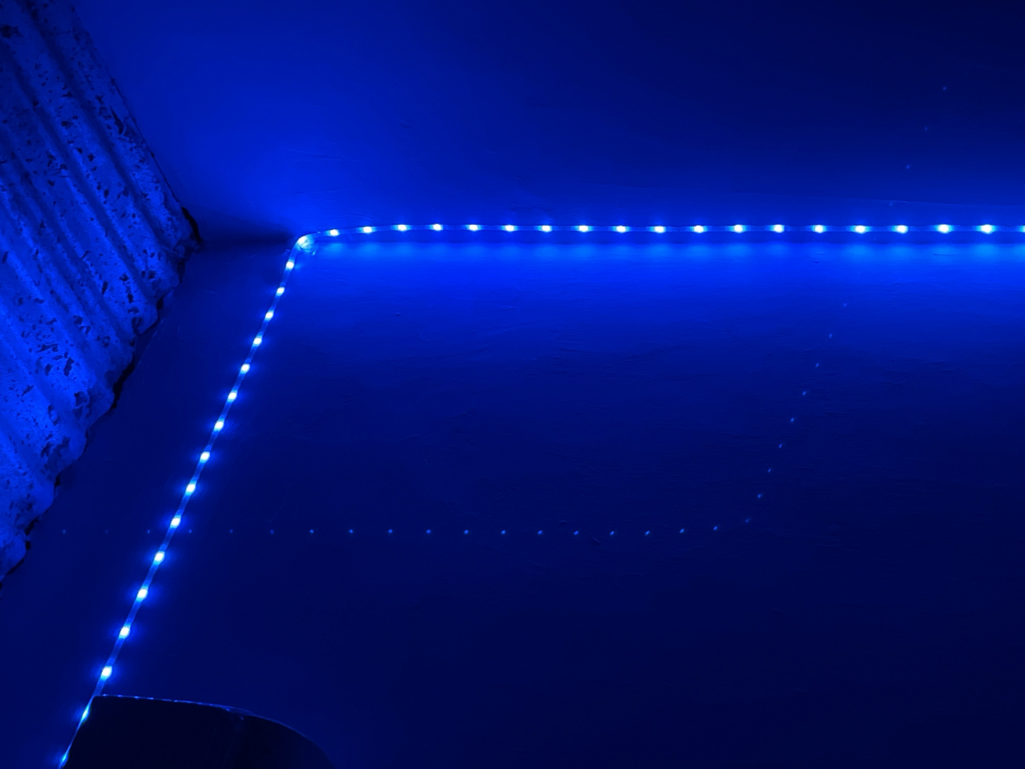 A low-angle, indoor shot depicts a section of a room illuminated by a string of blue LED lights. The lights are affixed along the corner where the wall meets the ceiling. The corner is also where the wall is next to a curtain. The light reflects downwards on the wall and there are small dots of light reflected on the wall. The shot does not include any people, and the location within Juan Griego, Venezuela, is not discernible from the provided image.