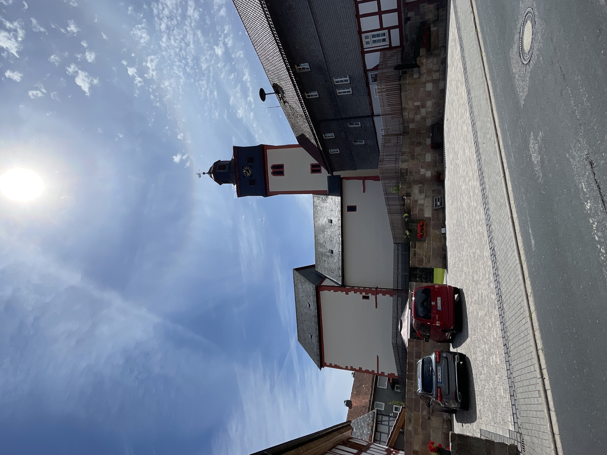 In Schwarzenborn, Germany, a town square on a sunny day features a tall church tower with a clock and a weather vane. The tower is adjacent to a large, white building with red trim, and further right, a dark-roofed building with traditional half-timbered elements. In the foreground, two parked vehicles, a gray SUV and a red van, are situated on a cobblestone paved area. Beyond the vehicles, a low stone wall separates the paved area from a raised grassy area with planters. The sky is blue with scattered white clouds and a bright sun.
