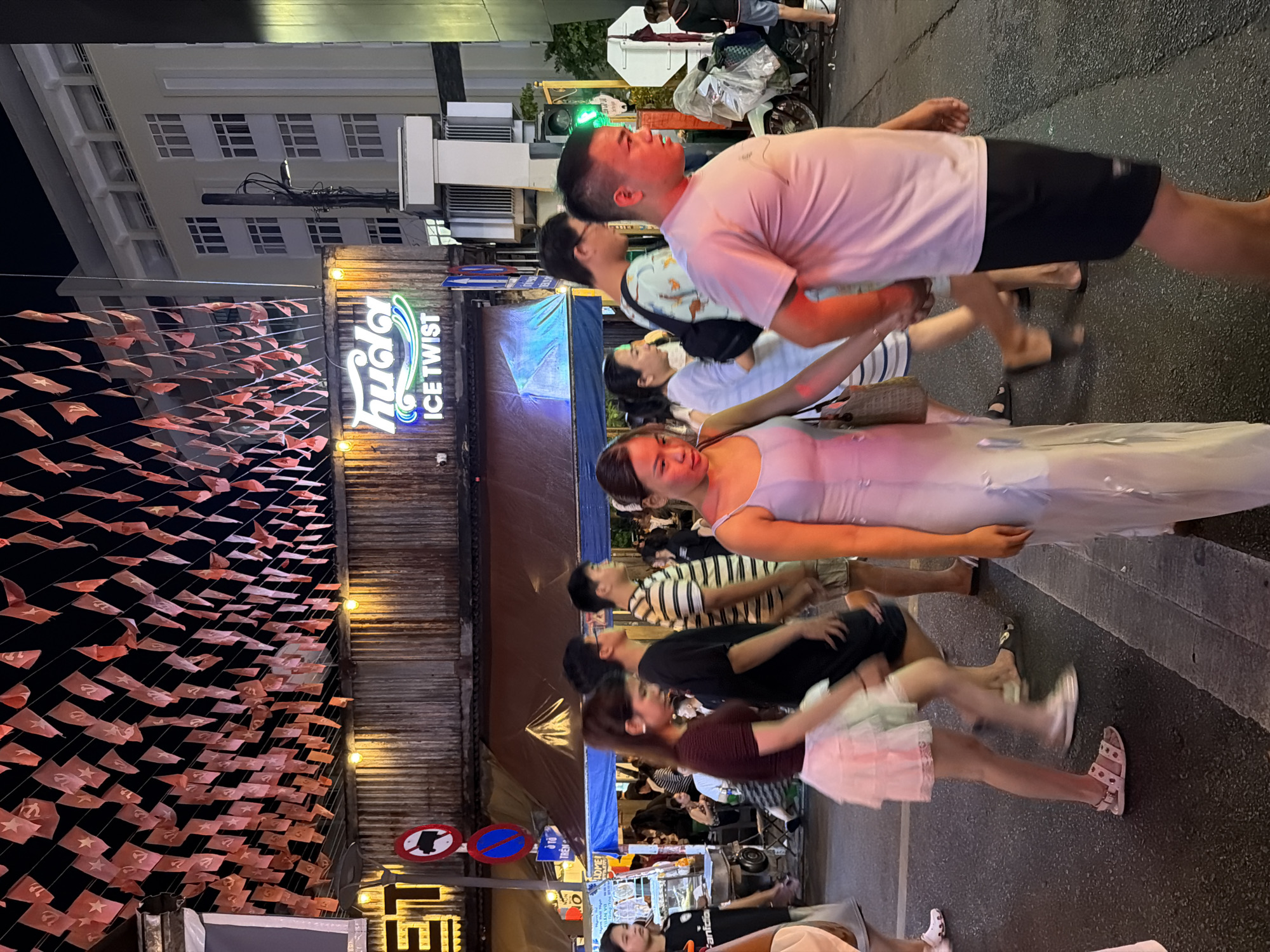 The image captures a street scene at night in Thuận Hóa Ward, Vietnam, characterized by a bustling atmosphere with pedestrians. Red flags with white stars and communist symbols are strung overhead, adorning the facade of a building that features a neon sign for "huda ICE TWIST." The street is paved, and a traffic light is visible. People are walking in various directions, some in groups and others individually. The individuals in the foreground are diverse in age and attire, with clothing ranging from casual t-shirts and shorts to dresses. Some people are carrying bags, and one person appears to be wearing sandals. In the background, a motorbike is parked, and a few more individuals are visible, contributing to the sense of a lively urban environment. The lighting suggests artificial illumination from storefronts and streetlights.