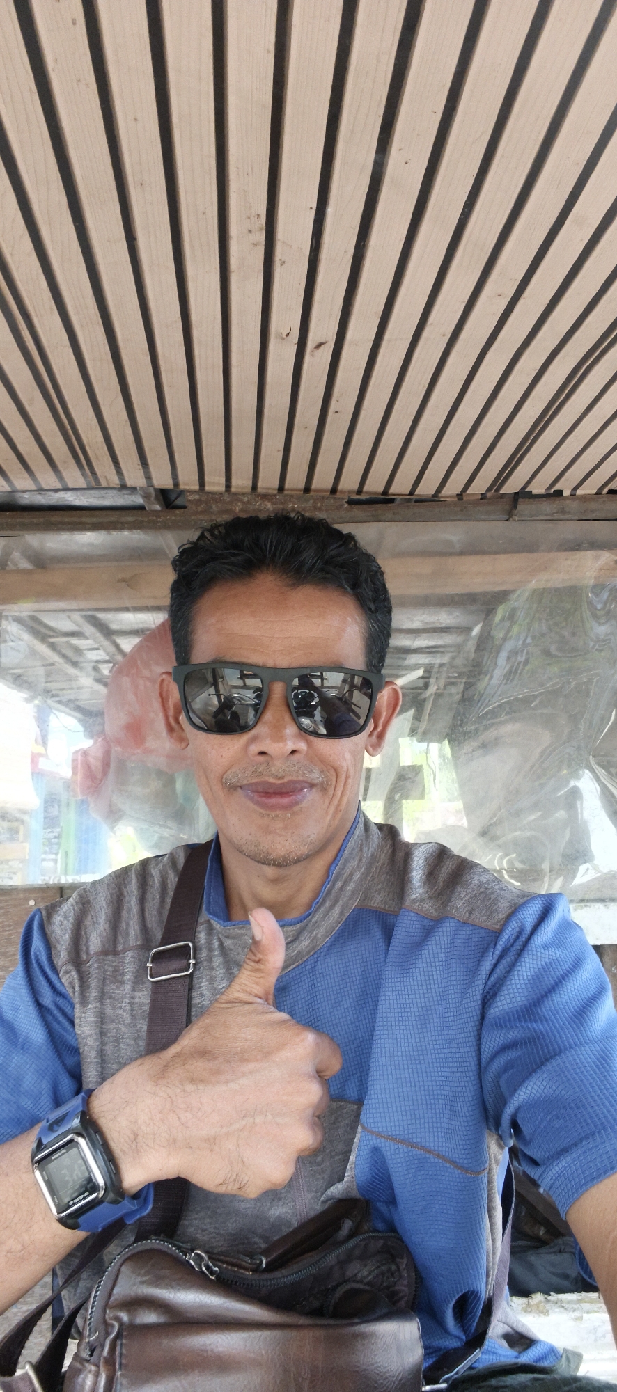 A medium shot captured from a slightly low angle shows a man wearing sunglasses and a blue and grey shirt. He is giving a thumbs-up gesture with his right hand and has a brown leather bag slung across his body. His reflection in the sunglasses shows a motorcycle. The background features a wooden slatted ceiling and a translucent barrier with some objects visible behind it. The scene appears to be outdoors, possibly in a market or roadside stall, common in areas like Muaro Jambi, Indonesia, given the man's attire and the ambient setting.