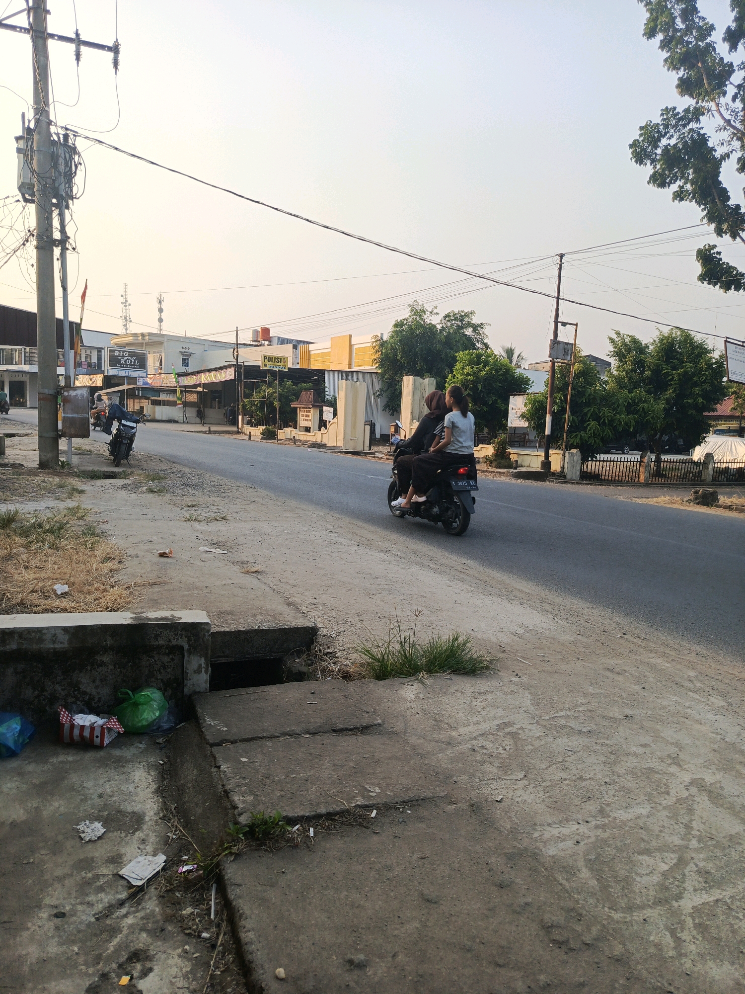 The image depicts a street scene in Talang Ubi, Indonesia, characterized by a clear sky and late afternoon sunlight. On the right side of the asphalt road, two individuals, both appearing to be female, are riding on a black scooter. The person in front is wearing a light-colored shirt and dark pants, while the person behind is wearing a dark top and dark pants. The scooter has a license plate with the characters "T 3075 RI".
To the left of the road, a concrete sidewalk and drainage ditch are visible. Litter is scattered on the ground near the ditch, including a green plastic bag, a red and white patterned box, and a blue plastic bag. A concrete culvert opening is present in the ditch. Further to the left, a utility pole with numerous wires and electrical equipment stands prominently.
In the background, several low-rise buildings are visible along the street, some with awnings. One building has a sign that reads "POLISI," indicating a police station. Trees and vegetation line the sides of the road and are interspersed among the buildings, suggesting a semi-urban or town setting. The overall impression is a common street scene in a populated area of Indonesia.