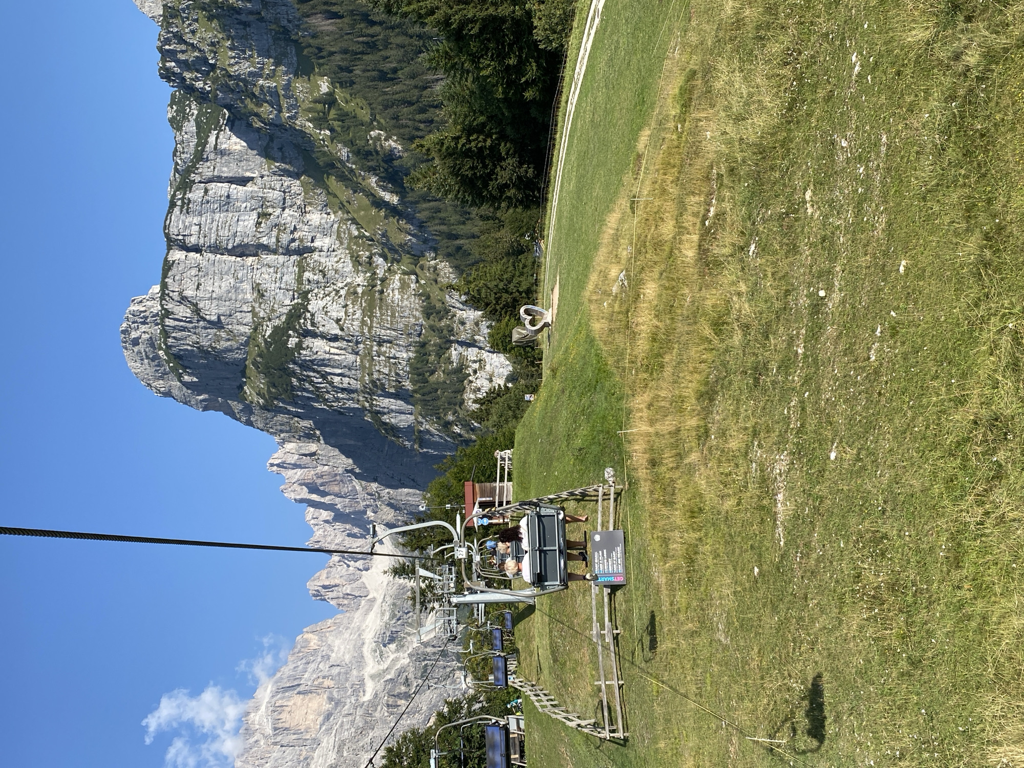 The image depicts a picturesque mountain landscape, likely in the vicinity of Molveno, Italy, given the context of the Dolomites. A ski lift with several occupied chairs ascends a grassy slope. In one chair, two individuals are seated, facing away from the camera. The chairs are suspended from a thick cable that stretches across the frame.
The environment is characterized by rolling green hills interspersed with patches of dry grass and scattered stones. Wooden fences line parts of the slope, and a dirt path is visible winding through the grass. In the background, towering, rugged mountains with steep, rocky cliffs dominate the skyline. Dense green pine forests cling to the lower slopes of the mountains. A large, abstract sculpture resembling a heart shape is situated on the grassy area to the right of the ski lift. The sky is a clear, bright blue with a few wispy white clouds. The overall scene suggests a sunny day in a mountainous region, likely during the summer or early autumn season.
