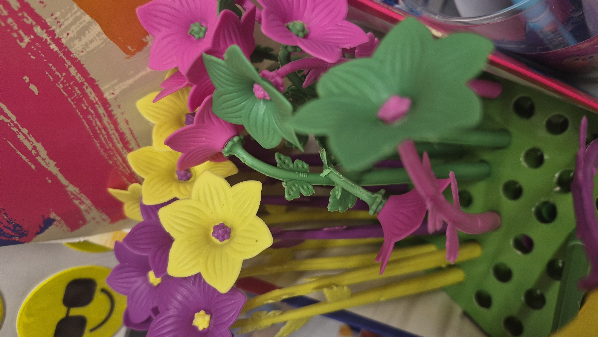 The image shows a close-up of a display of colorful plastic flowers on thin stems, arranged in a green perforated stand. The flowers are in various shades of purple, yellow, and pink, with some green leaves and stems also visible. In the background, there is a blurry pattern of pink, white, and orange, possibly part of a packaging or display. To the left of the flowers, a portion of a yellow circular sticker with sunglasses and a smiley face is visible. The scene appears to be an indoor display, likely in a store or market, but there is no identifiable information linking it to Jēkabpils, Latvia. No people are present in the image.