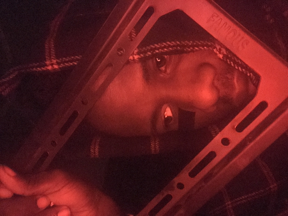 A person's face appears through a square metal frame with the word "FAMOUS" inscribed. The person wears a head covering with a checkered pattern. The lighting is dim and predominately red, casting the image in a monochromatic hue. The person's hand can be seen in the lower-left corner. A gold tooth is visible in the person's mouth. The composition is close-up and angled.