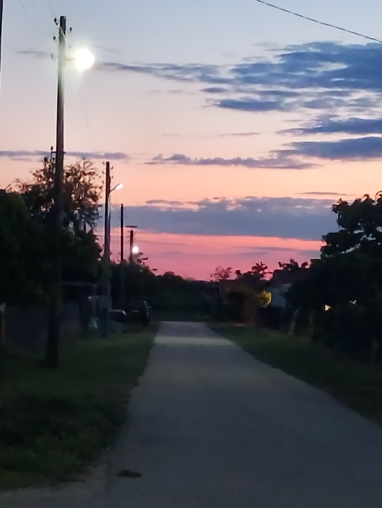 The image depicts a rural road at dusk, with a soft pink and blue sky transitioning into night. A single, unpaved road extends into the distance, flanked by dark green grass and silhouetted trees. Several utility poles with visible wires are spaced along the sides of the road, with one pole featuring a bright, illuminated streetlamp. The sky is partially covered by scattered, dark clouds, contrasting with the lighter hues of the sunset. The overall impression is one of peaceful solitude in a rural setting.