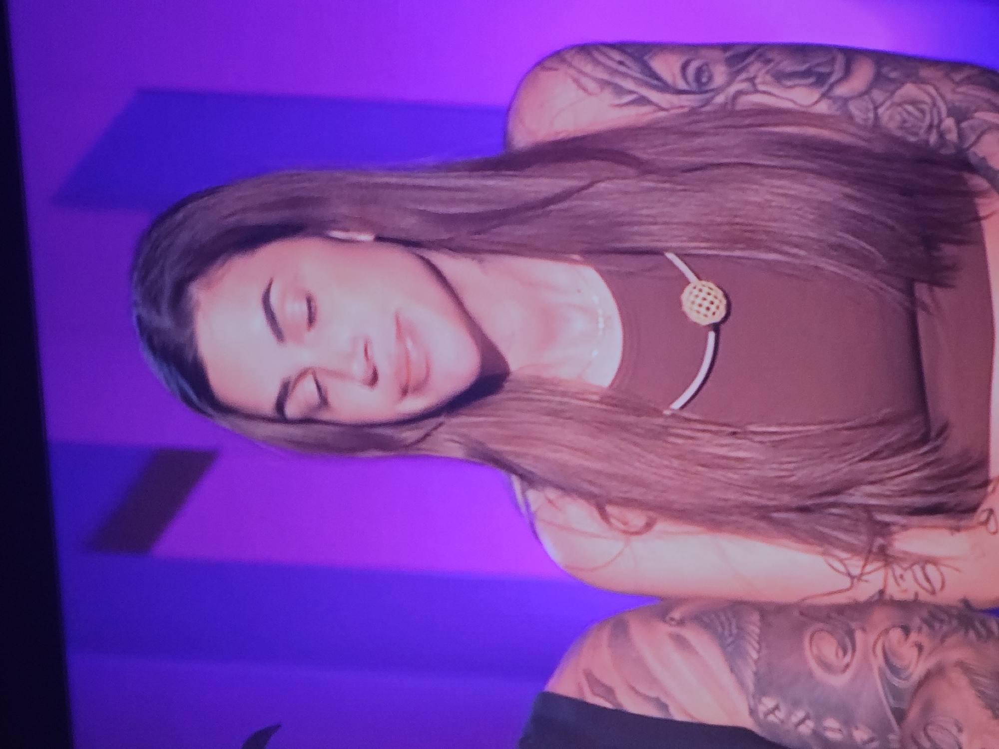 The image shows a close-up of a woman with long, dark brown hair and multiple tattoos on her arms. She is wearing a dark brown, sleeveless top and a thin necklace with a gold, circular pendant. The background is a vibrant purple color with some indistinct darker shapes. The woman's eyes are closed, and she has a slight smile. To her left, a portion of another person's arm, also heavily tattooed, is visible. The scene appears to be indoors, possibly a television studio or a similar controlled environment. There are no discernible elements in the image that can place the location within the city of Buchen (Odenwald), Germany.