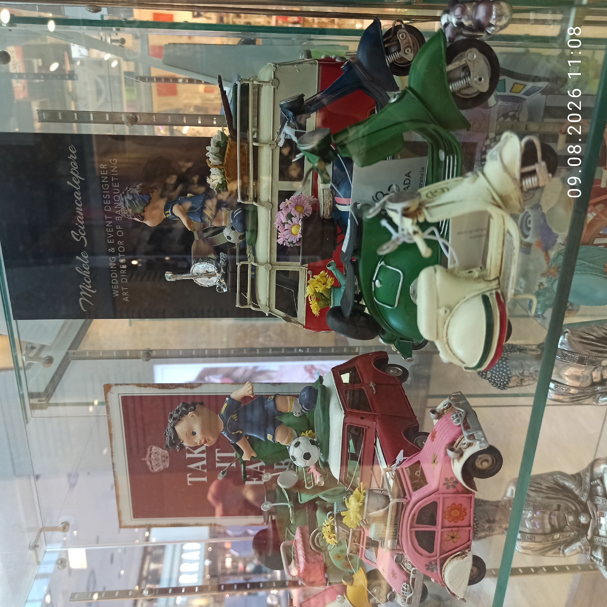 The image is a medium shot of a display case in a retail setting, likely a gift shop or a store specializing in miniatures. The focus is on various miniature vehicles and figurines arranged within the glass enclosure. In the foreground, a cream-colored miniature scooter is prominent, with a green scooter partially visible behind it. To the left, a pink miniature car with floral decorations is displayed above a silver statue of Buddha. Behind these, a red miniature car is visible, and further back, a red and white miniature van is positioned with figurines of two children and miniature musical instruments on its roof rack. A sign with text including "Michele Sciancalepore" and "WEDDING & EVENT DESIGNER" is visible in the background, reflecting in the glass. The overall scene is brightly lit, suggesting an indoor environment. The bottom right corner of the image shows a timestamp, "09.08.2026 11:08". There is no indication of the specific location within Molfetta, Italy, beyond it being a commercial display.