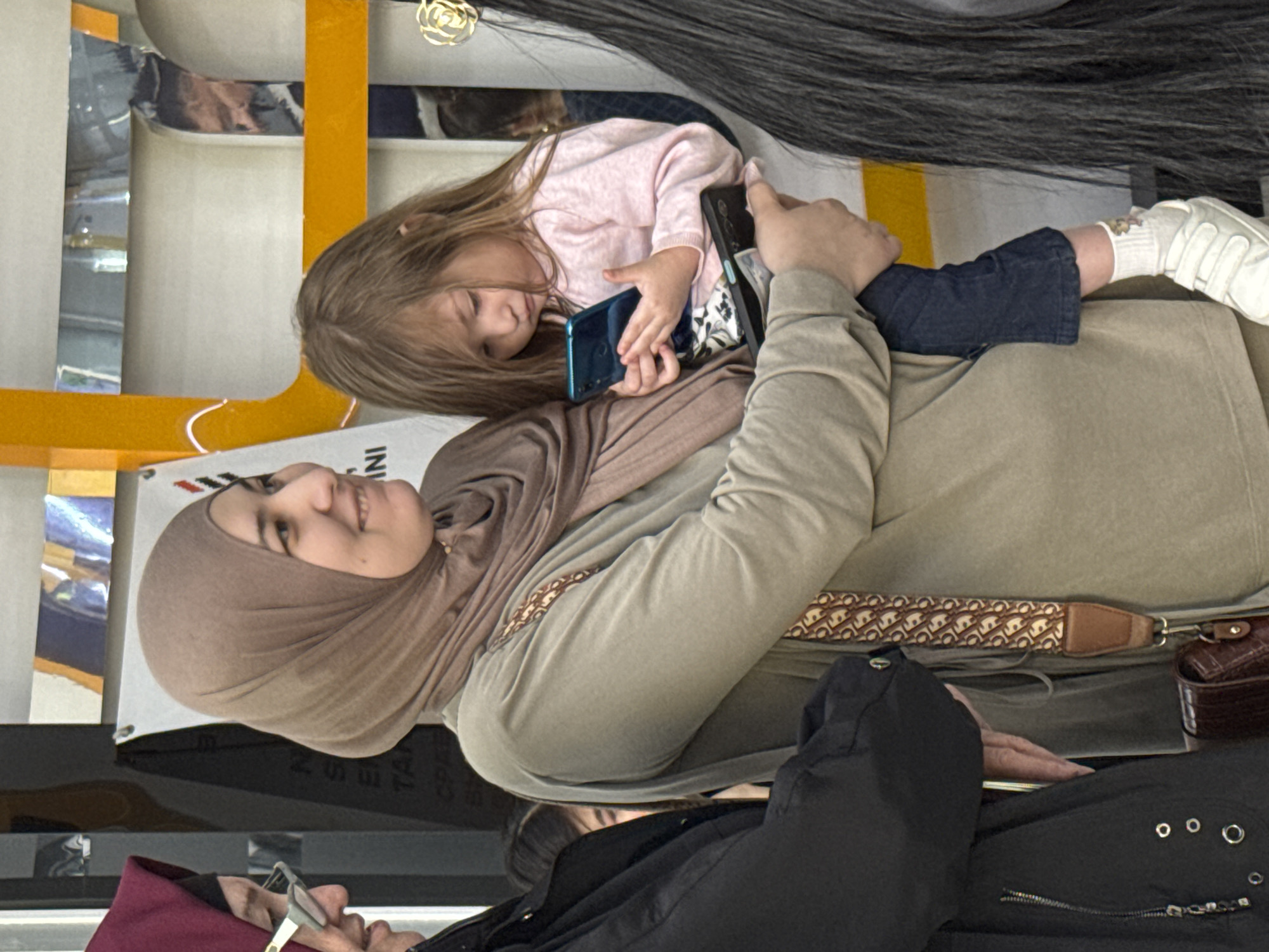 The image shows a woman wearing a hijab sitting down and holding a young girl on her lap. The girl is holding a blue smartphone, while the woman holds a black smartphone. Both are looking at the phones. The woman is wearing a light brown hijab and a matching long-sleeved shirt. The girl is wearing a light pink sweater and blue jeans with white shoes. The woman has a purse strap with a repeating patterned design. The scene takes place inside what appears to be a shopping mall or a similar indoor public space. In the bottom of the image an older adult is visible. The location might be Tashkent, Uzbekistan, based on contextual clues.