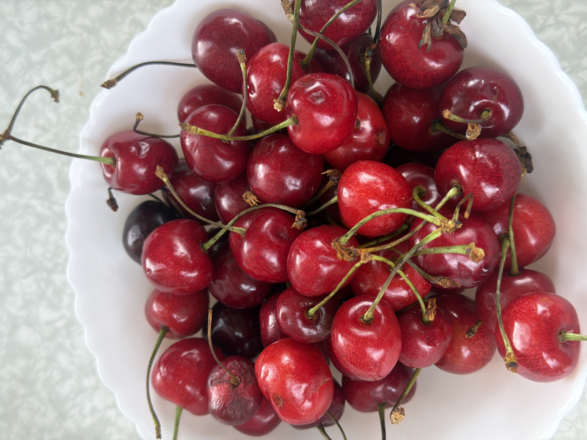 A close-up, overhead shot shows a bowl filled with fresh, ripe cherries. The cherries are a deep, glossy red, with some appearing almost black. They are of varying sizes and shapes, with some having their stems still attached, while others are stemless. The bowl itself is white and has a ruffled edge, typical of a serving dish. The cherries are piled high, creating a rich texture and a vibrant display of color. The background is a light-colored, textured surface, possibly a tablecloth or placemat. The lighting is soft and natural, suggesting it was taken during the daytime. There are no people or any other objects visible in the image. The overall impression is one of abundance and freshness, highlighting the natural beauty of the fruit.