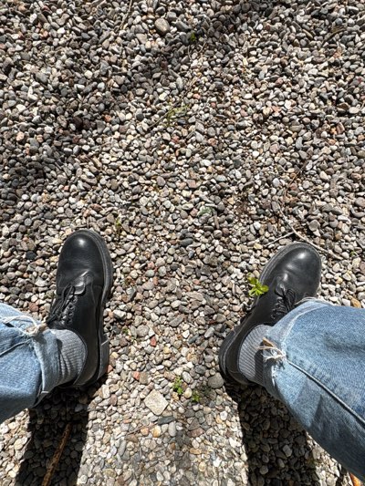 An overhead view depicts the lower extremities of an individual resting on a gravel surface in Erba, Italy. The ground consists of numerous small, irregularly shaped stones in shades of gray, white, and brown, with scattered sparse green vegetation and dried organic matter. Distinct shadows are cast across the gravel, indicating direct sunlight. The individual is wearing light blue denim jeans, which exhibit minor fraying at the cuffs, revealing light gray ribbed socks. Both feet are shod in black, low-heeled, lace-up shoes with thick, dark soles, positioned slightly apart and stationary on the gravel.