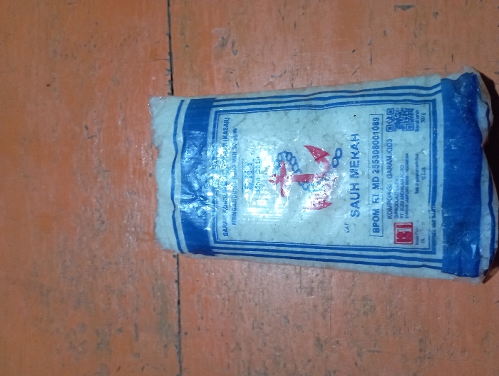 The image displays a white and blue plastic package of salt resting on an aged, orange-brown wooden surface. The package, slightly crumpled, contains coarse iodized salt.

Visible text on the package includes:
- Product type: "GARAM KONSUMSI BERYODIUM (KASAR)" (Iodized Consumer Salt (Coarse))
- Iodine content: "MENGANDUNG IODIUM MIN 30 PPM" (Contains minimum 30 ppm Iodine)
- Brand name: "CAP SAUH MEKAH" (Brand: SAUH MEKAH), accompanied by a logo featuring a red anchor and a white infinity symbol.
- BPOM registration number: "BPOM RI MD 255308001089" (Indonesian Food and Drug Authority).
- Composition: "KOMPOSISI: GARAM, KIO3" (Composition: Salt, Potassium Iodate).
- Producer: "DIPRODUKSI OLEH PT. DUDI ANDALAN AGRO" (Produced by PT. Dudi Andalan Agro).
- Producer location: "PUNGGUR LAMPUNG INDONESIA".
- Net weight: "BERAT BERSIH 300 g".
- A QR code is also visible on the package.

The wooden surface shows distinct plank lines and some wear, suggesting an indoor setting. There is no activity depicted; the image is a still life of the packaged product. The lighting is diffused, consistent with an indoor environment. The location context provided is Pubian, Indonesia, which aligns with the product's origin in Lampung, Indonesia.