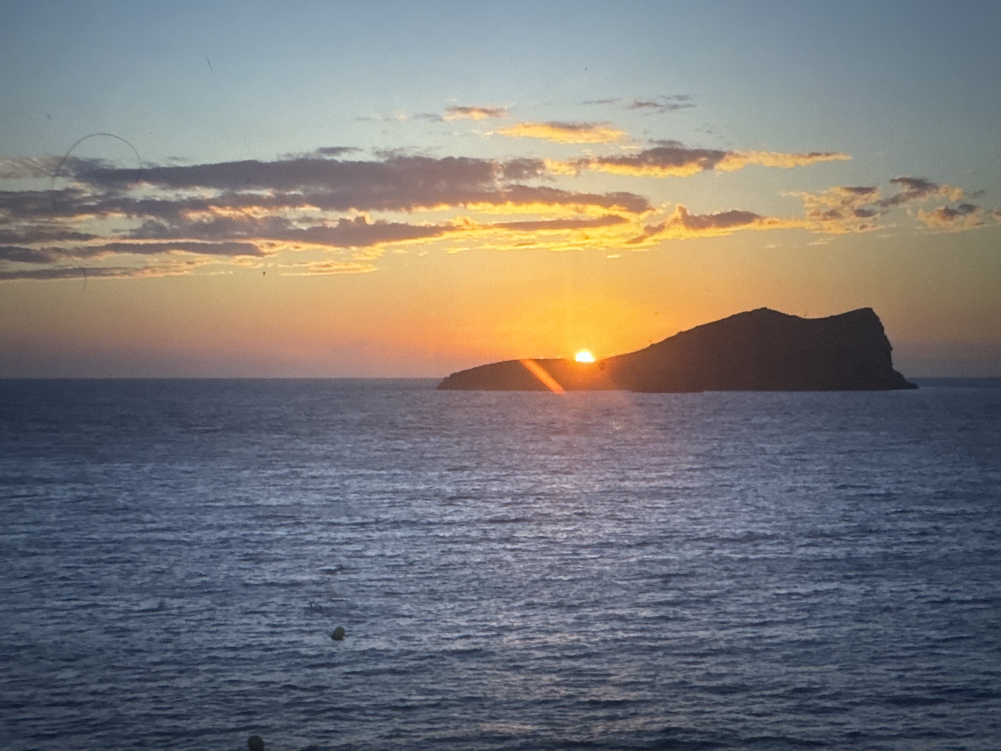 A serene ocean sunset is depicted, with the sun partially obscured by a silhouetted island. The sky transitions from a pale blue at the top to a warm orange hue near the horizon, where several clouds are illuminated by the setting sun. The ocean is a deep blue, with gentle ripples on its surface. There are a few small, dark buoys visible on the water. The image does not contain any elements that suggest it is located in Porrentruy, Switzerland, as it portrays a coastal or island landscape.