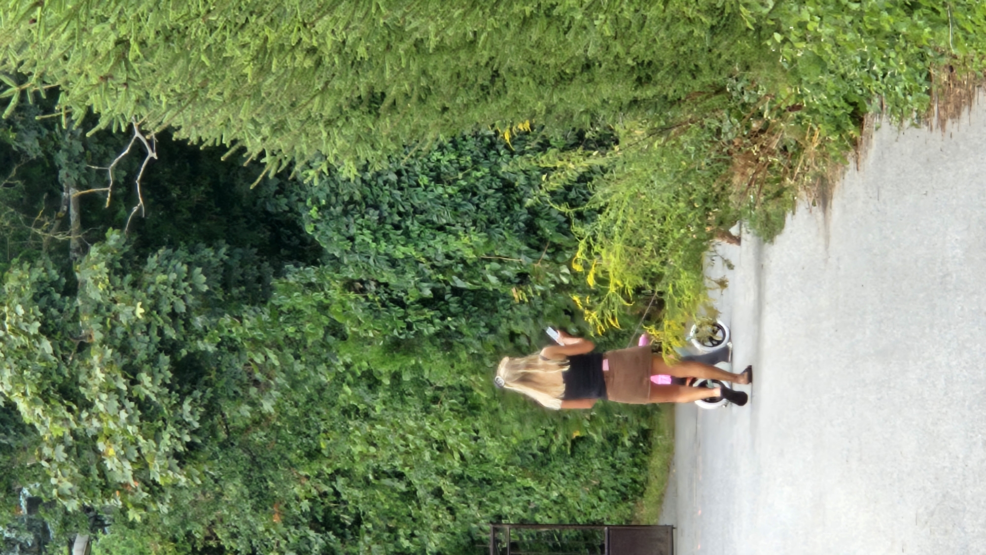 The image depicts a blonde woman from behind, walking on a paved path. She is wearing a black top and a brown skirt. She is holding a mobile phone in her right hand and appears to be looking at it. Behind her, a child is on a pink tricycle with white wheels. They are walking beside a dense green hedge that forms a wall on the left side of the path. To the right of the path, there is lush green vegetation, including tall trees. The scene appears to be in an outdoor setting, possibly a park or a residential area, with a fence visible on the far left. The specific location in Prešov, Slovakia, is not identifiable from the image alone.