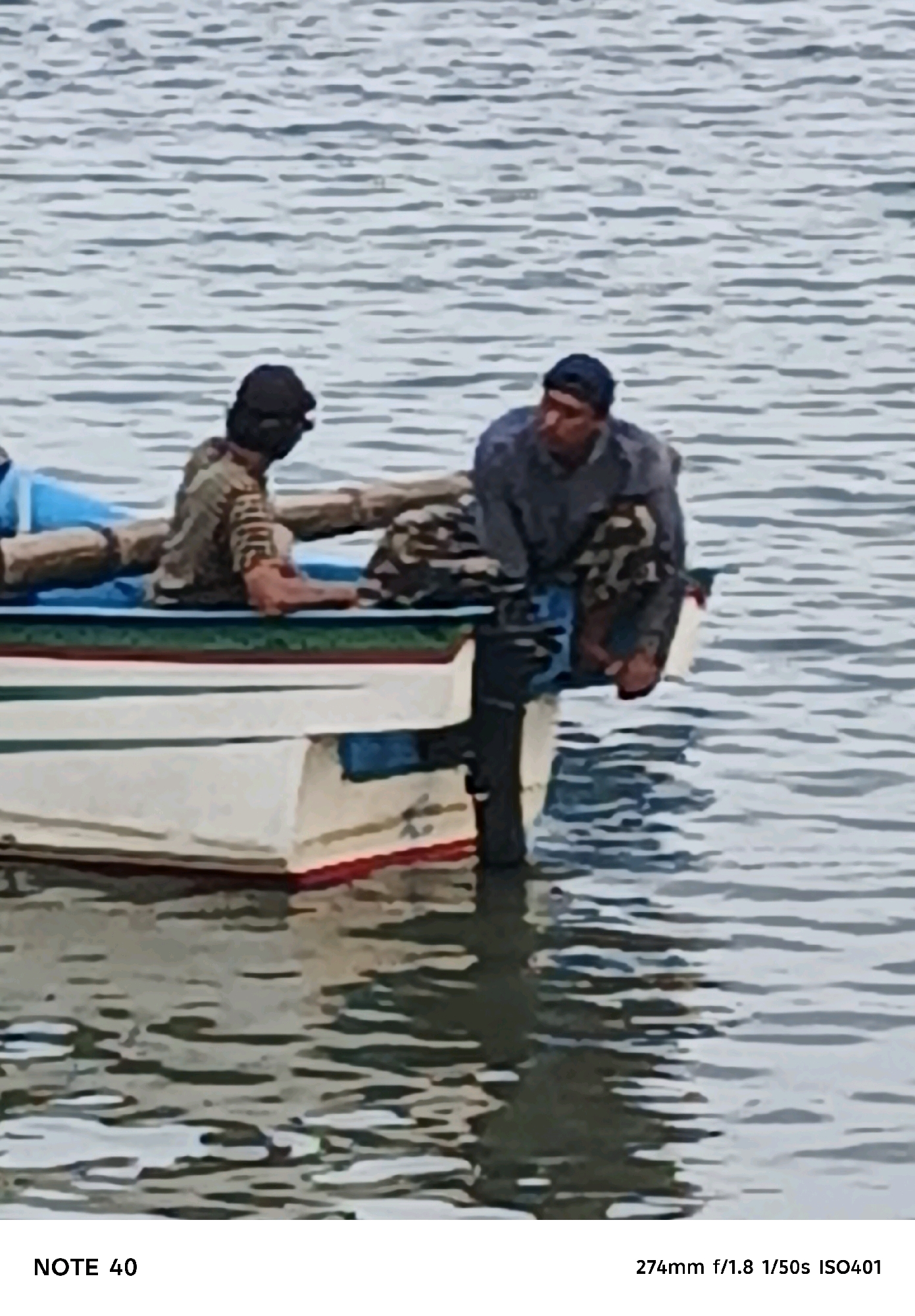 Two individuals are seated in a small boat on rippling water. The person on the left, wearing a dark cap and a camouflage-patterned shirt, is looking towards the right. The person on the right, wearing a dark blue cap and a grey long-sleeved shirt, is looking down. The boat is white with red and green trim around the upper edge. The water is a muted blue-grey. The scene is likely located on the coast of Kelurahan Tegalkamulyan, Cilacap, Indonesia.