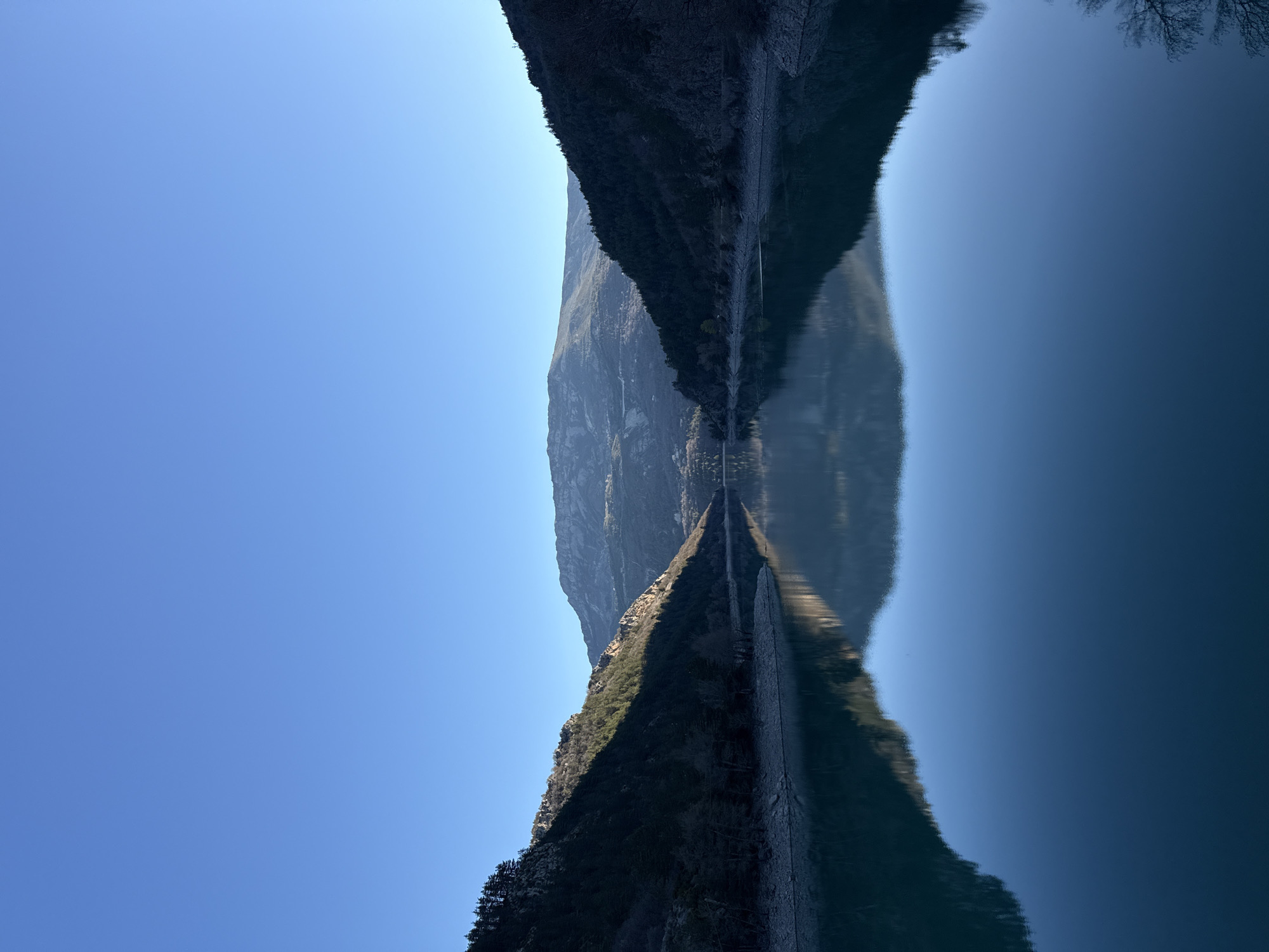 The image depicts a body of water, likely a lake, reflecting a mountain range under a clear blue sky. The mountains are textured with visible rock formations and some vegetation. The reflection is almost perfect, creating a symmetrical composition. The location is likely near Castellane, France, given the terrain and general landscape. No people, subjects, objects, actions, or interactions are visible beyond the natural landscape.
