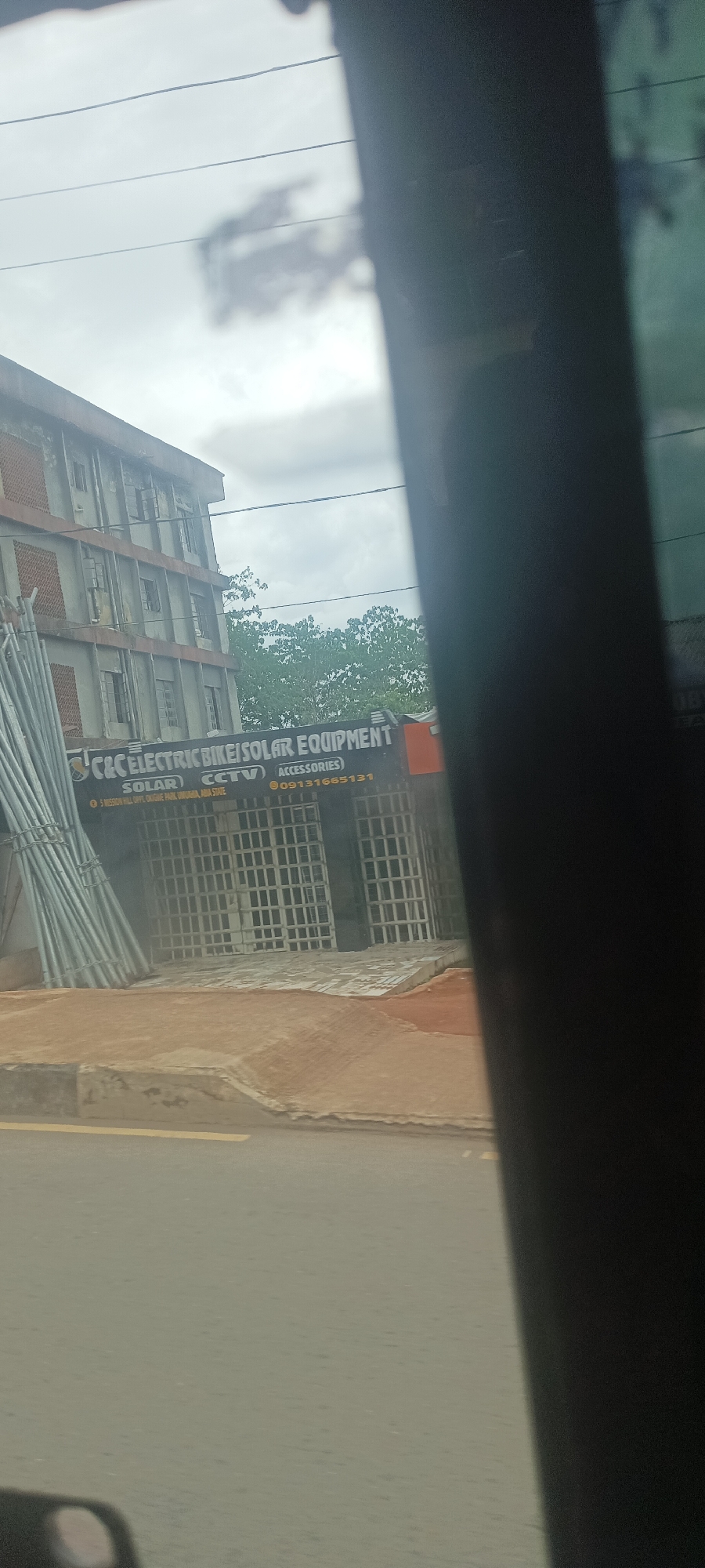The image, taken from within a vehicle, shows the exterior of a business named "C&C Electric Bike/Solar Equipment". The business provides services related to solar power, CCTV, and bike accessories, and is located at 5 Mission Hill, opposite Okigwe Park, in Umuahia, Abia State, Nigeria. The phone number for the business is 09131665131.
The building appears to be a multi-story structure with what seem to be closed window coverings. A pile of metal poles or pipes rests on the left side of the building. The business has metal security bars in front of the storefront. The image is somewhat obscured by a dark object in the foreground, most likely part of the vehicle interior. The sky appears overcast. A road runs in front of the building.