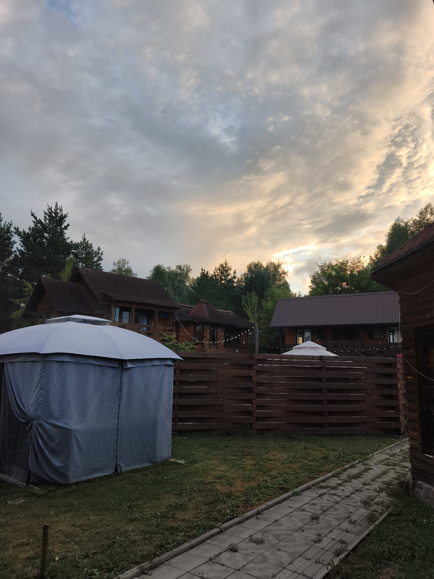 In Катунь, Russia, this outdoor scene depicts a tranquil setting with a cloudy sky and a soft glow from the setting sun. In the foreground, a gray gazebo with a white roof is set up on a grassy area. Behind it, a rustic wooden fence separates the yard from a line of wooden houses nestled amongst lush green trees. The houses appear to be vacation cabins or guest houses, with their warm, natural wood exteriors and dark brown roofs. String lights are visible on one of the houses, suggesting it might be evening or a festive occasion. A paved walkway runs alongside the fence, leading further into the property. The overall atmosphere is peaceful and inviting, typical of a rural or resort-like environment.