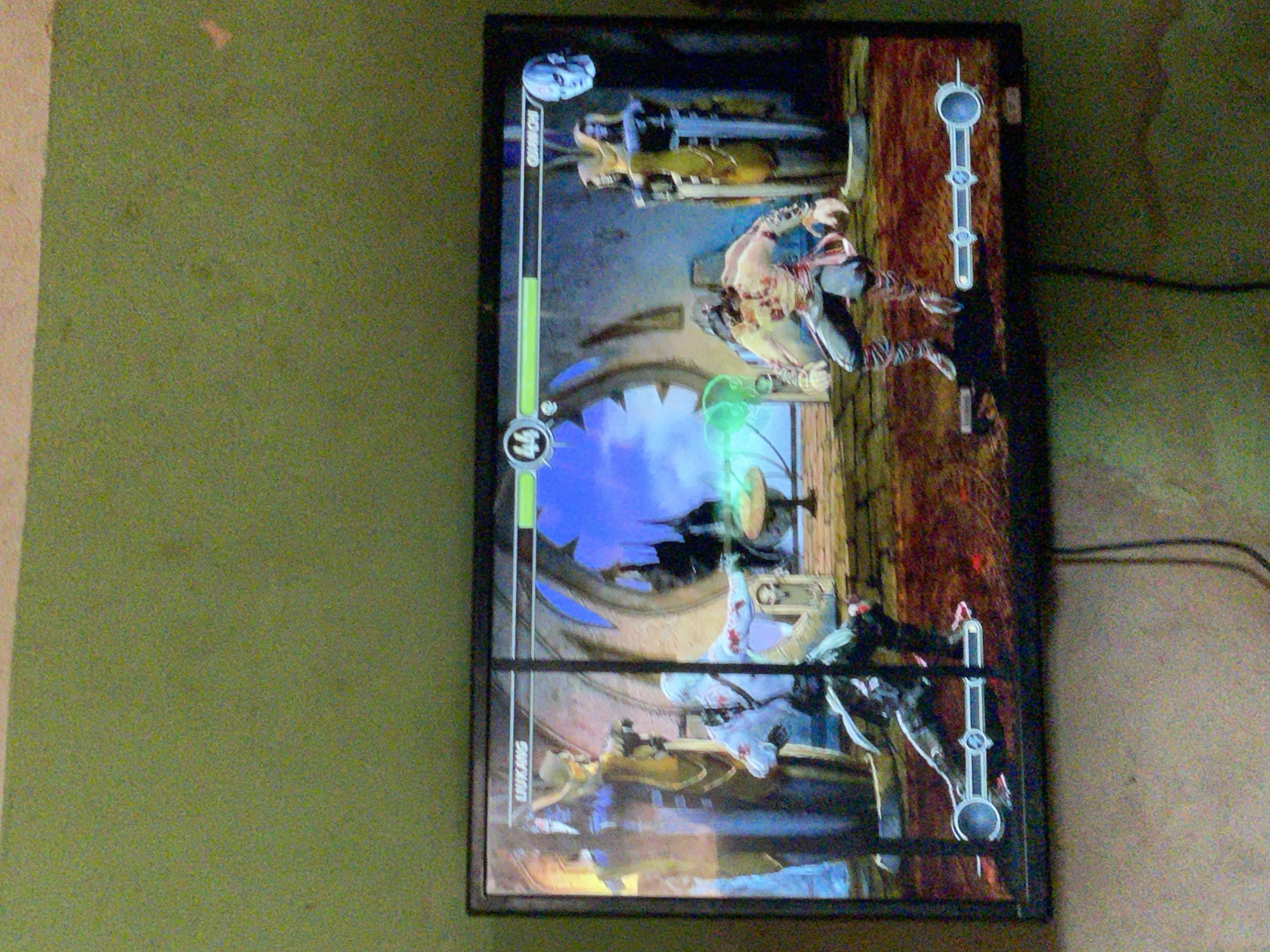 A television screen displays a video game, seemingly a fighting game, likely Mortal Kombat. Two characters are in combat, with visible health bars above them labeled "Bo' Rai Cho" and "Liu Kang". The game stage features a gothic-style archway and a stone platform. The screen is mounted on a wall, positioned vertically. The wall to the left of the screen appears to be painted a greenish hue, while the wall to the right is a speckled beige. Two black cables extend from the right side of the television. The location is Akwanga, Nigeria.