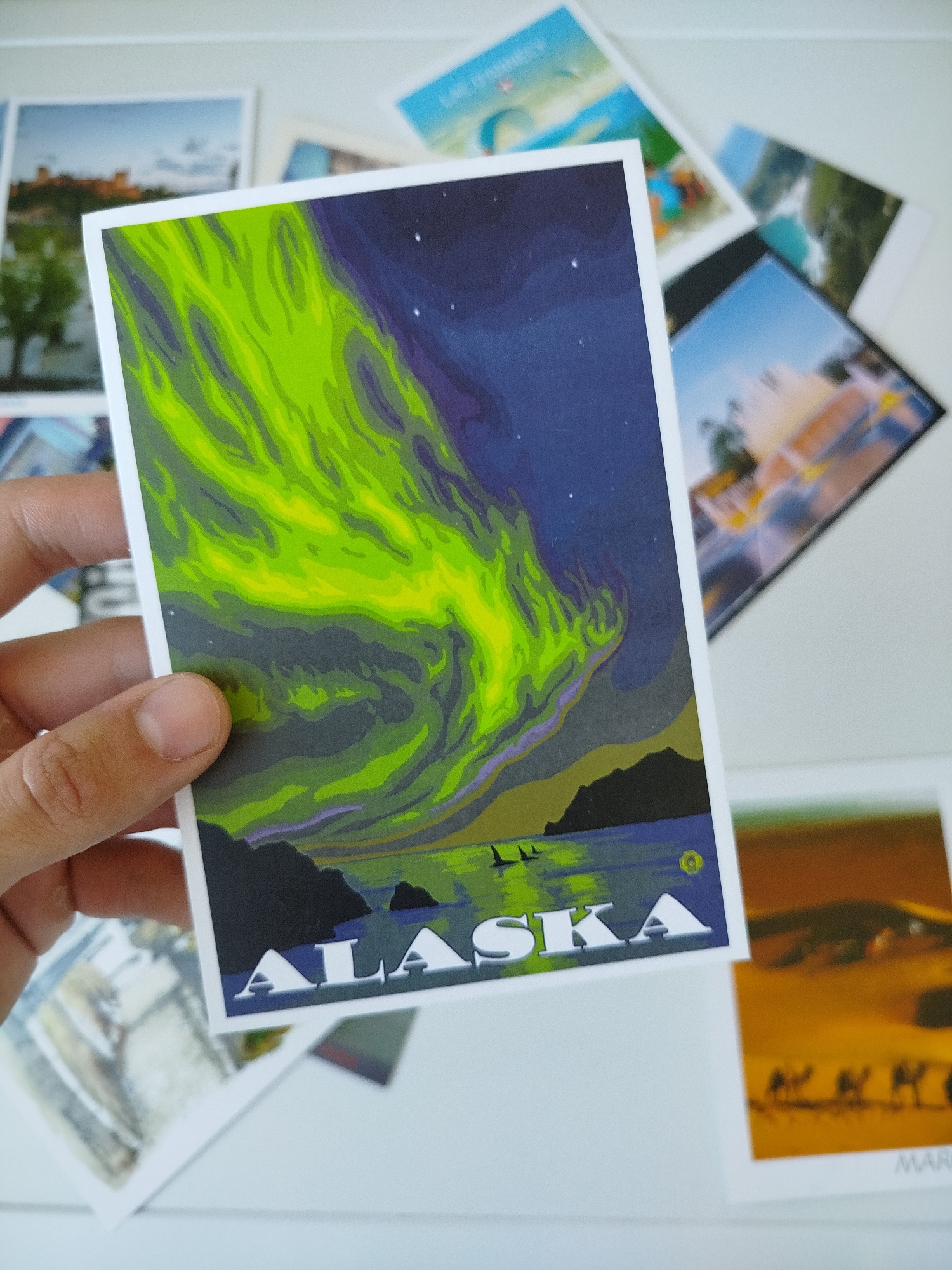 A person holds a postcard featuring an illustration of the Aurora Borealis above water with landmasses in the foreground. The word "ALASKA" is printed in white letters at the bottom of the postcard. Other postcards are scattered around the main card. The image does not contain any information relating to Varna, Bulgaria.