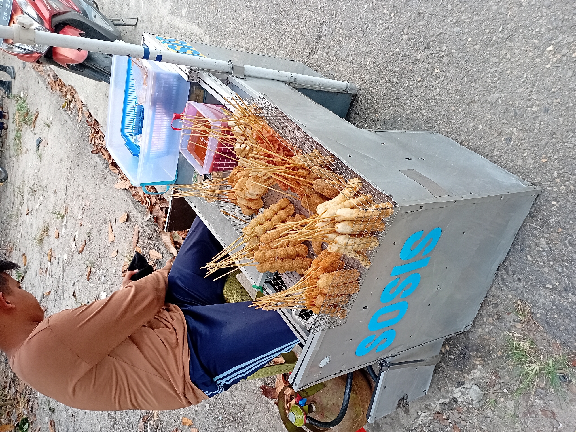 A street vendor in Jambi, Indonesia, operates a mobile cart selling deep-fried sausages and meatballs on skewers. The vendor, wearing a brown long-sleeved shirt and navy blue track pants with white stripes, is seated and engaged with a black animal, possibly a cat, on his lap. The cart is constructed of grey metal and features the word "SOSIS" in large blue lettering. Displayed prominently on the cart are several wire baskets filled with various deep-fried items on wooden skewers, ready for sale. A red plastic container filled with a red sauce, likely chili sauce, is also visible. To the left of the cart sits a gas cylinder, a common sight for food vendors. In the background, a red motorcycle is parked, and the ground is a mixture of dirt, gravel, and scattered dry leaves. The overall environment suggests an outdoor, likely roadside, setting in Jambi, Indonesia.