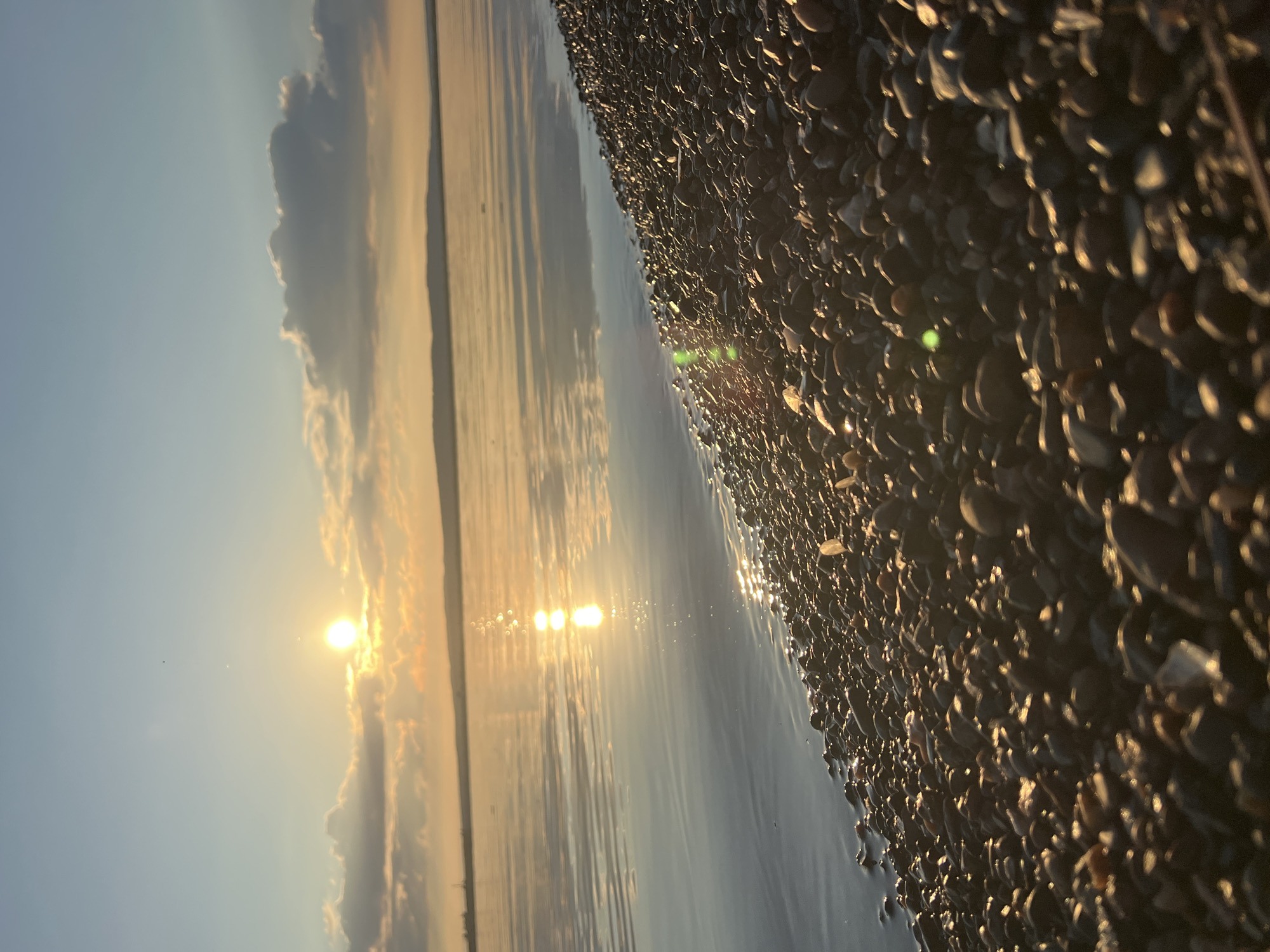 The image captures a tranquil beach scene during sunset in Chaikovsky, Russia. The sun is low on the horizon, casting a warm golden glow across the water and the shore. The sky is a clear blue with a few wispy clouds tinged with orange and yellow hues. The sun's reflection shimmers on the calm surface of the water, creating a mesmerizing visual effect.
The foreground is dominated by a rocky beach, with a multitude of smooth, dark-colored pebbles. The pebbles are wet from the receding tide, reflecting the sunlight and adding a subtle sparkle to the scene. The water gently laps at the edge of the beach, creating a sense of peace and serenity. The overall mood of the image is one of quiet beauty and natural splendor, evoking feelings of calm and relaxation. The scene suggests a peaceful end to the day, with the natural beauty of the coastline bathed in the soft light of the setting sun.