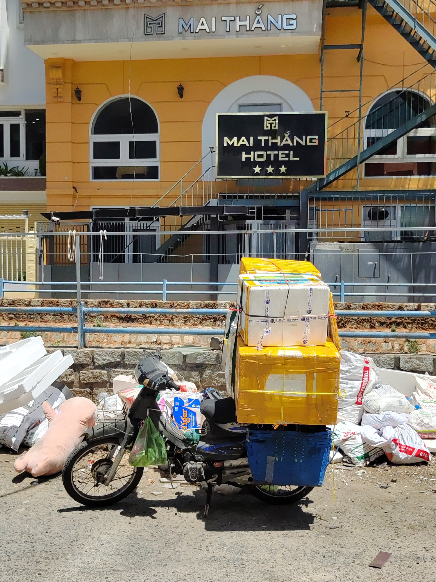 The image captures a parked motorbike heavily loaded with packages in front of the Mai Thắng Hotel in Da Lat, Vietnam. The motorbike has a blue basket attached to the rear, and the packages are wrapped in yellow tape and white boxes. Debris and bags, including a stuffed pink object, are scattered around the motorbike on the roadside. The Mai Thắng Hotel, an orange building, is visible in the background with its sign and architectural details, including arched windows and a staircase. A protective blue railing is set between the street and the hotel, partially obscured by the debris. The street appears to be in an urban setting.