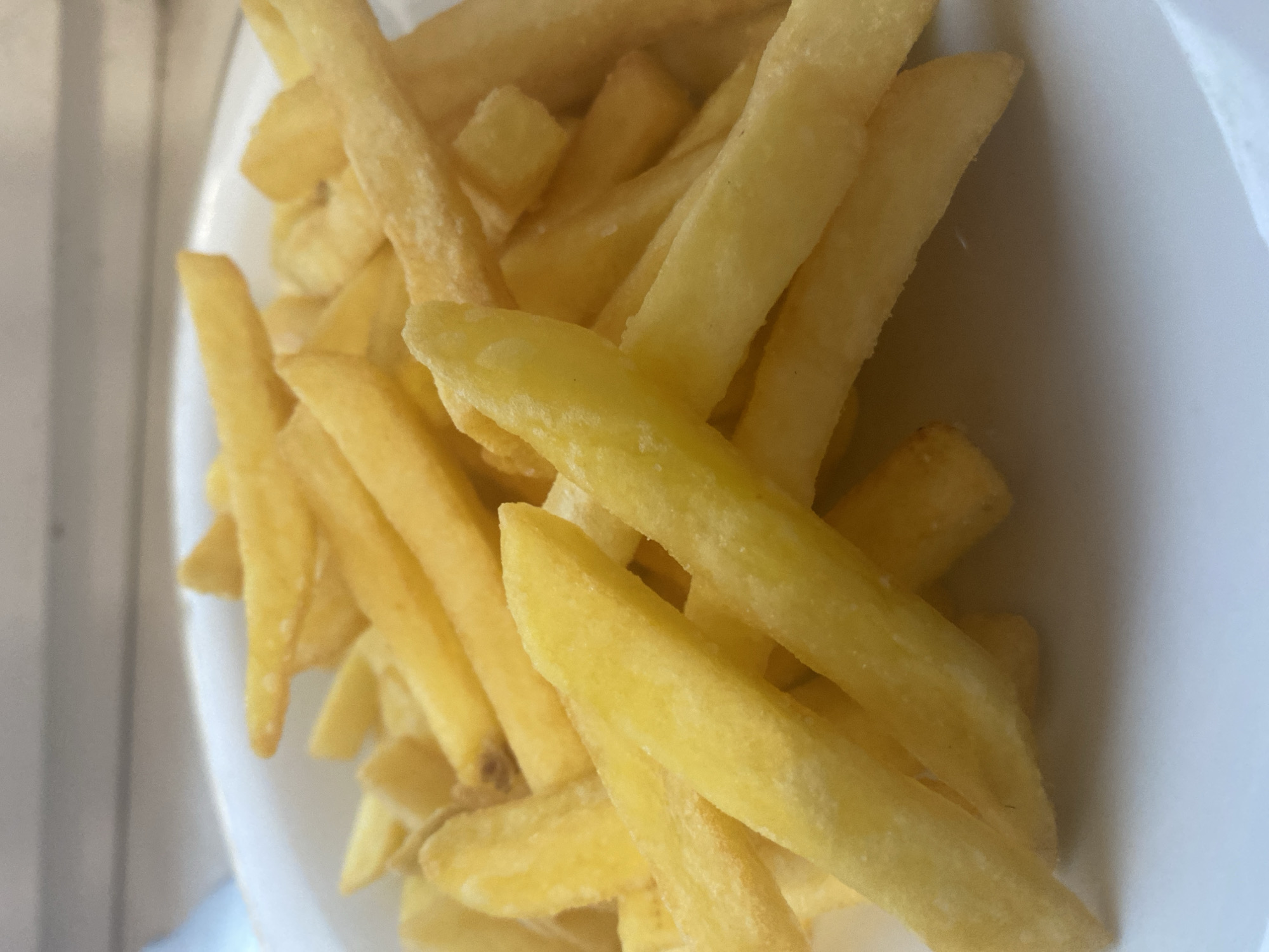 A close-up shot features a white plate piled high with golden-brown french fries. The fries are cut into classic shoestring shapes and are piled in a mound, with some overlapping others. The lighting casts shadows and highlights on the fries, emphasizing their texture and crispiness. The background is blurred, suggesting the focus is entirely on the food. The setting appears to be an indoor environment, possibly a restaurant or home, given the plate and the subdued background. There are no people or animals visible in the image.