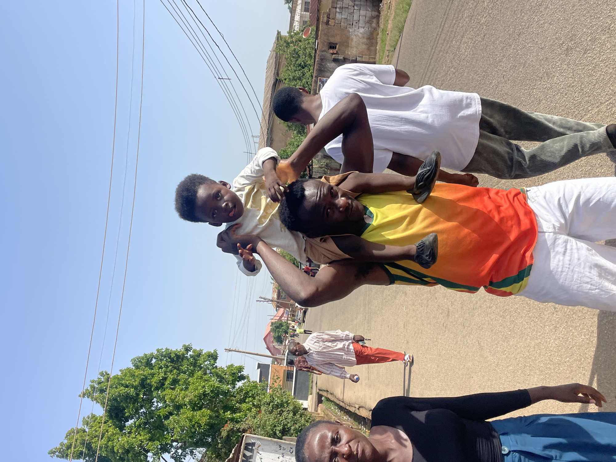 The image shows a group of people on a street in Jos, Nigeria. A man in a yellow, orange, and green shirt and white pants is carrying a young boy in a light-colored shirt and khaki shorts on his shoulders. Another man in a white shirt and olive green corduroy pants is walking behind them. In the foreground, a woman in a black top and blue pants is visible. Other people are walking in the background. The sky is clear and blue, and there are buildings and trees along the street. Electrical wires are visible overhead. The ground appears to be paved with asphalt.