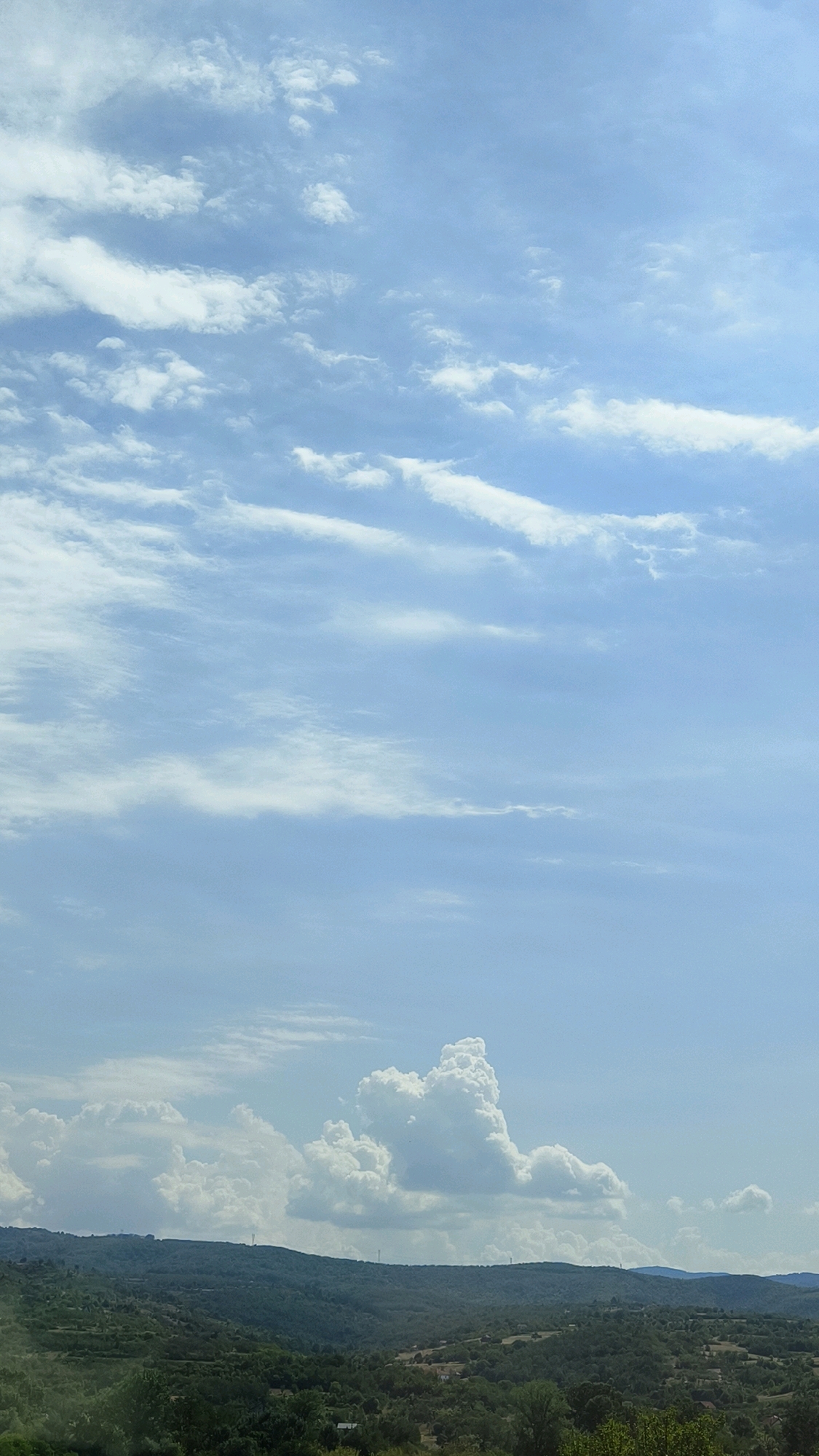 A photograph captures a hazy, expansive sky filled with scattered white clouds. The clouds range from wispy, horizontal streaks to larger, cumulus formations that cast subtle shadows. Below the sky, a rolling, green landscape unfolds, characterized by rolling hills covered in dense vegetation and scattered trees. The terrain appears to be agricultural, with visible terraces and possibly small structures or dwellings nestled within the landscape. In the distance, several tall, slender towers, likely telecommunications or wind turbines, rise above the horizon, breaking the natural silhouette of the hills. The overall impression is one of a rural, possibly Mediterranean or Balkan, environment under a bright, overcast sky. The location is identified as Vranje, Serbia.