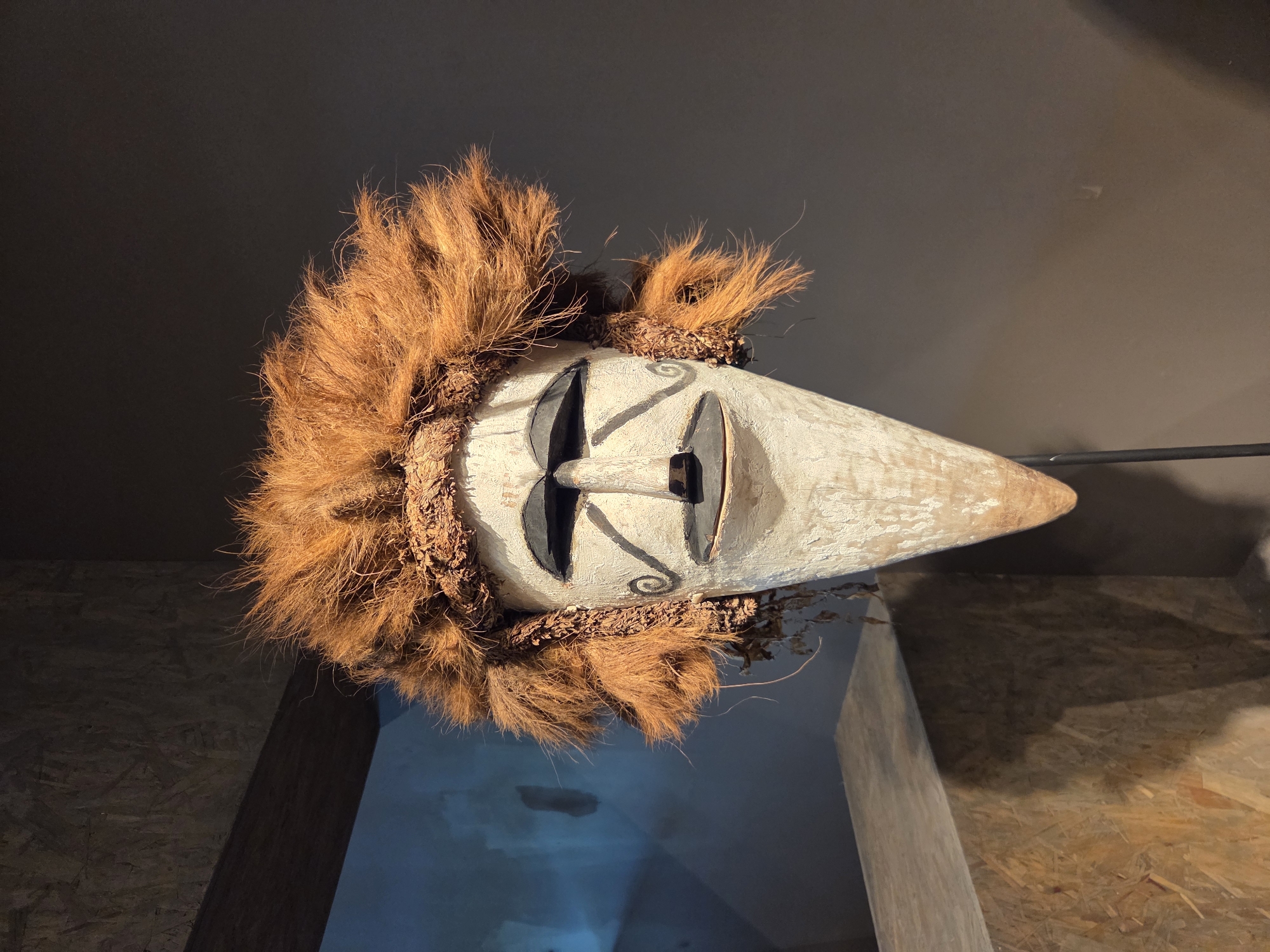 This is a medium shot, eye-level, indoor photograph of a tribal mask displayed in a museum or gallery setting. The mask is the primary subject, positioned centrally and angled slightly to the right. It is made of wood and painted white, with black painted features for the eyes, nose, and mouth. The eyes are large and almond-shaped with thick black outlines. The nose is a simple, dark rectangle. The mouth is open and also appears as a dark, rectangular shape. The mask is adorned with a headdress made of shaggy, reddish-brown natural fibers, resembling hair or fur. There are also smaller pieces of woven material and dried plant matter attached around the sides of the mask, near where ears might be. The mask has a long, conical shape, tapering to a point at the bottom.
The mask is mounted on a thin, black metal stand, which is not very visible in the frame. Behind the mask, the background transitions from a darker, textured wall on the right to a lighter, possibly blue-tinted wall on the left. Above the mask, there is a wooden beam or shelf, part of the museum's architectural structure. In the lower left of the image, a portion of a wooden display structure, possibly a pedestal or bench, made of particleboard or OSB, is visible. The lighting in the scene is dramatic, with the mask well-lit, highlighting its features and texture, while the background is darker.
The image was taken at the Bowers Museum in Santa Ana, California, United States of America. The mask itself is identified as a "ceremonial mask" from Papua New Guinea.