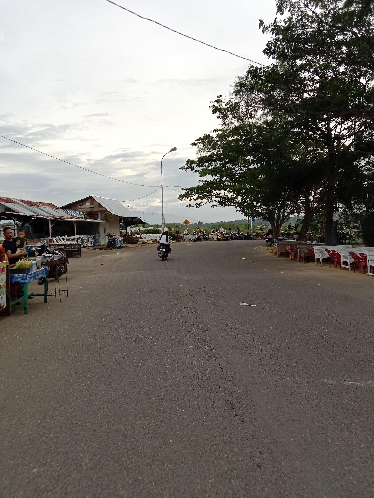The image depicts a street scene in Lhokseumawe, Indonesia. The paved road stretches into the distance, where it forks around a cluster of trees. A motorcyclist is traveling along the road towards the fork. To the left of the road are several simple structures with corrugated metal roofs, possibly shops or stalls. A man is standing with a young child next to a stall that has food items. To the right of the road, there are rows of red and white plastic chairs and benches under the trees. Further in the distance is a gathering of people and parked motorcycles. Overhead, utility lines stretch across the sky. The sky is overcast with clouds.
