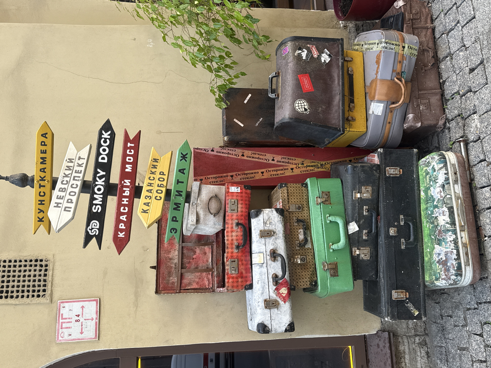 The image displays a quaint exterior scene featuring a wall adorned with colorful directional signs pointing to various Saint Petersburg landmarks, including "KUNSTKAMERA," "NEVSKY PROSPECT," "SMOKY DOCK," "KRASNY MOST," "KAZANSKY SOBOR," and "ERMITAGE." Below these signs, a collection of vintage suitcases is stacked against the wall. The suitcases vary in size, color, and material, with some being leather and others fabric-covered. Several of the suitcases are adorned with stickers, and one is wrapped with tape labeled in Russian "Осторожно стекло!" meaning "Caution glass!". A small, white square sign with red markings is also visible on the wall, possibly indicating a map or a route. The overall setting appears to be a street or an alleyway, with cobblestone paving visible on the right side of the image. Lush green foliage cascades from above, adding a touch of nature to the urban landscape. The scene has a nostalgic and artistic feel, suggesting a creative display or a themed establishment. The lighting suggests it is daytime, with no specific weather conditions immediately apparent. No people are visible in the frame.