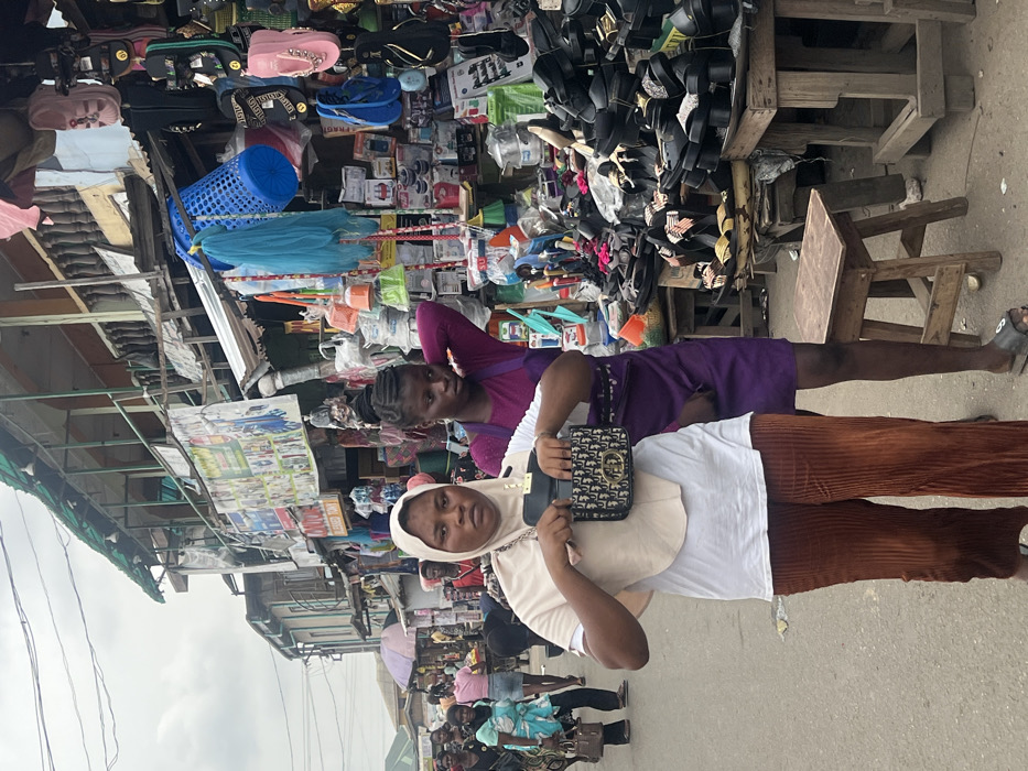 The image depicts a bustling outdoor market street in Egan, Nigeria, under an overcast sky. In the foreground, a person wearing a light beige hijab over a white t-shirt and brown corduroy pants holds open a small, dark handbag featuring a gold-colored "CD" style logo, looking directly at the camera with a neutral expression. To their right and slightly behind, another individual with braided hair, wearing a solid purple dress or apron-style garment, stands near a market stall, their left hand touching the back of their head. They wear a single dark slide sandal on their visible foot.
The background is densely packed with market stalls and activity. To the immediate right, a wooden stall displays numerous shoes, sandals, and flip-flops, predominantly black, stacked and arranged on trays and wooden structures. Above this, household items such as plastic buckets, brushes, and containers are displayed, some hanging, alongside brightly colored flip-flops and a blue plastic laundry basket. Further into the background, multiple multi-story buildings line the street, featuring weathered facades, exposed wooden balconies, and corrugated metal awnings. Various signs, including one partially visible with "BLESSED TOWN," are affixed to these structures. Numerous other individuals are visible throughout the street, walking and engaging in market activities. The street pavement is a light gray. Overhead, utility lines crisscross the sky. Two small wooden stools rest on the pavement in the foreground.