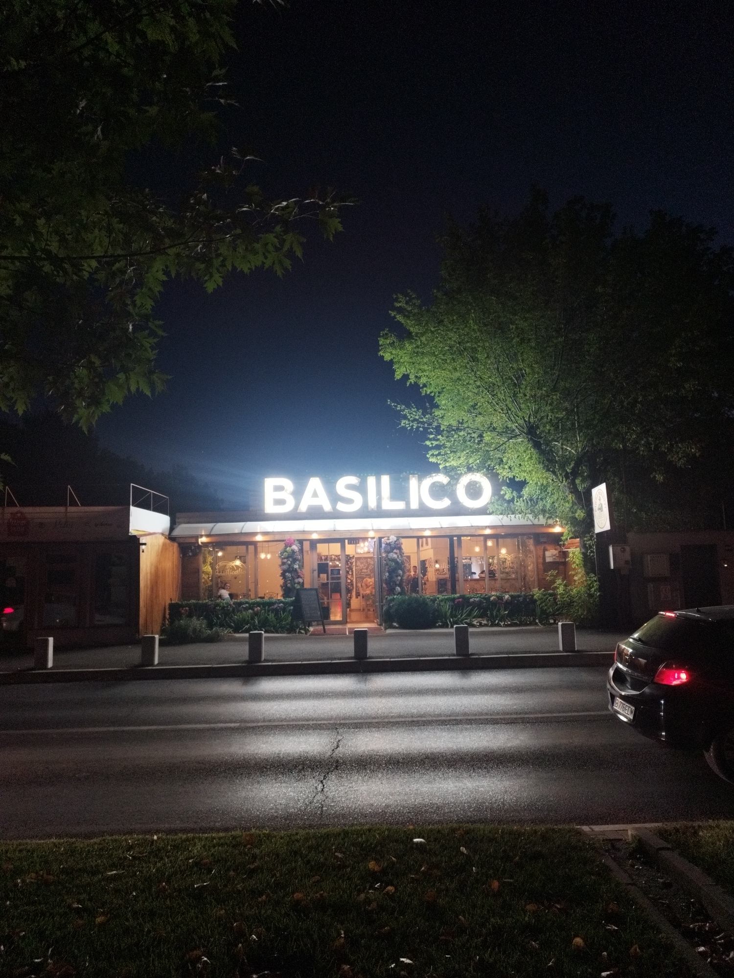 This is a nighttime, eye-level, wide shot of the restaurant "BASILICO" located in Bucharest, Romania. The restaurant's name is illuminated by large, white, capital letters above its entrance. The building has large windows, revealing a well-lit interior with patrons seated at tables. Green foliage surrounds the restaurant, and a large tree is visible to the right of the building. In the foreground, a dark-colored car is parked on the right side of a wet, asphalt road, its brake lights illuminated. The road appears to be an urban street, and the scene is illuminated by artificial lighting from the restaurant and the car.