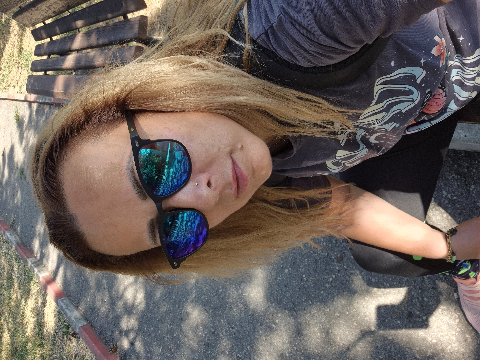 A medium shot captures a woman wearing sunglasses and a graphic t-shirt, seated outdoors on a sunny day. She has blonde hair, a nose piercing, and her left arm is resting on her lap. The background shows a paved path, a wooden bench, and grassy areas. The woman's sunglasses reflect a scene with trees and a fence. Her t-shirt has a colorful, abstract design. A shadow of a phone is visible on the ground near her feet, suggesting she may be taking a selfie.