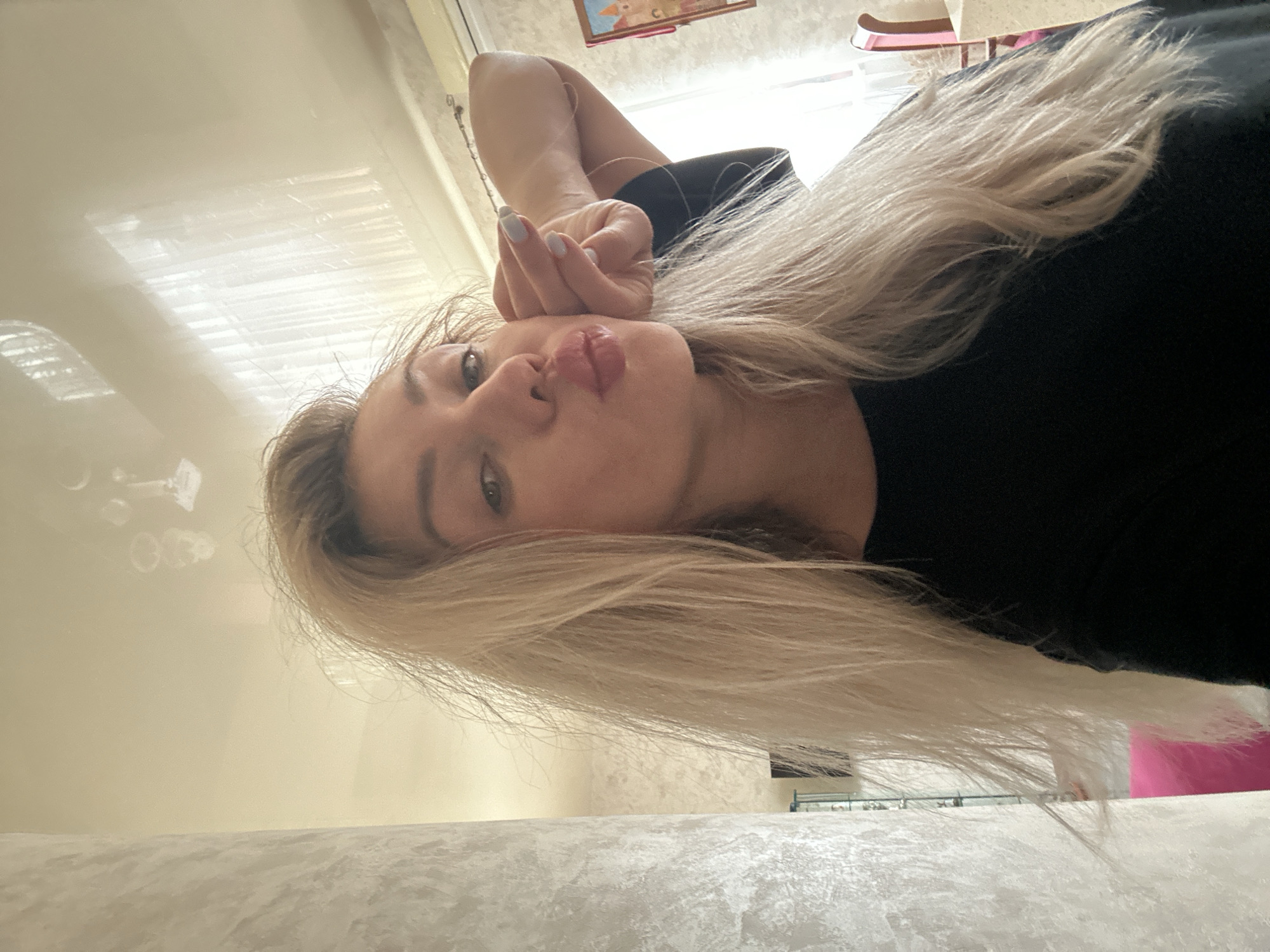 A young woman with blonde hair pulled back from her face is posing for a selfie. She is wearing a black shirt and has her left hand touching her cheek, with her lips puckered. The background appears to be an indoor setting, possibly a home or hotel room, with a textured wall on the left and a window with blinds on the right. There are reflections on the ceiling, suggesting it is a smooth, possibly glossy surface. The overall lighting is soft and diffused. The image does not contain any specific geographical markers that would indicate the location is Armavir, Russia.