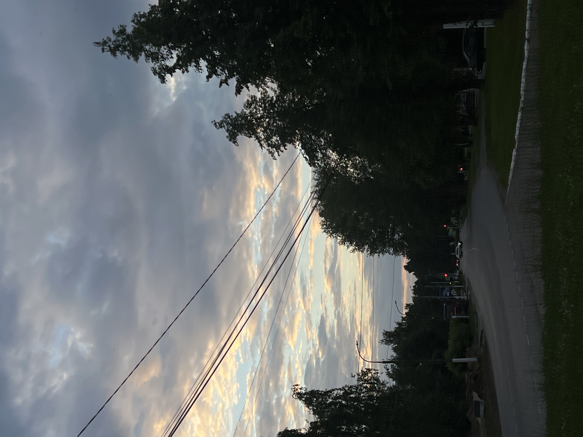 The image depicts a rural road scene in Chaikovsky, Russia, at dusk. The sky is overcast with dramatic clouds, tinged with the warm colors of sunset near the horizon. Several power lines crisscross the sky from left to right. Silhouetted trees line both sides of the road, with some appearing to be deciduous and others more coniferous.
On the left side of the road, a red truck is parked near a pile of dirt and construction materials. Further down the road, a white car is visible, driving away from the camera. Traffic lights are present, with at least one showing a red light. The road itself appears to be asphalt and curves gently to the right. On the right side of the road, a grassy area is visible, bordered by a white curb. The overall impression is one of a quiet, somewhat isolated, semi-urban or rural setting as daylight fades.