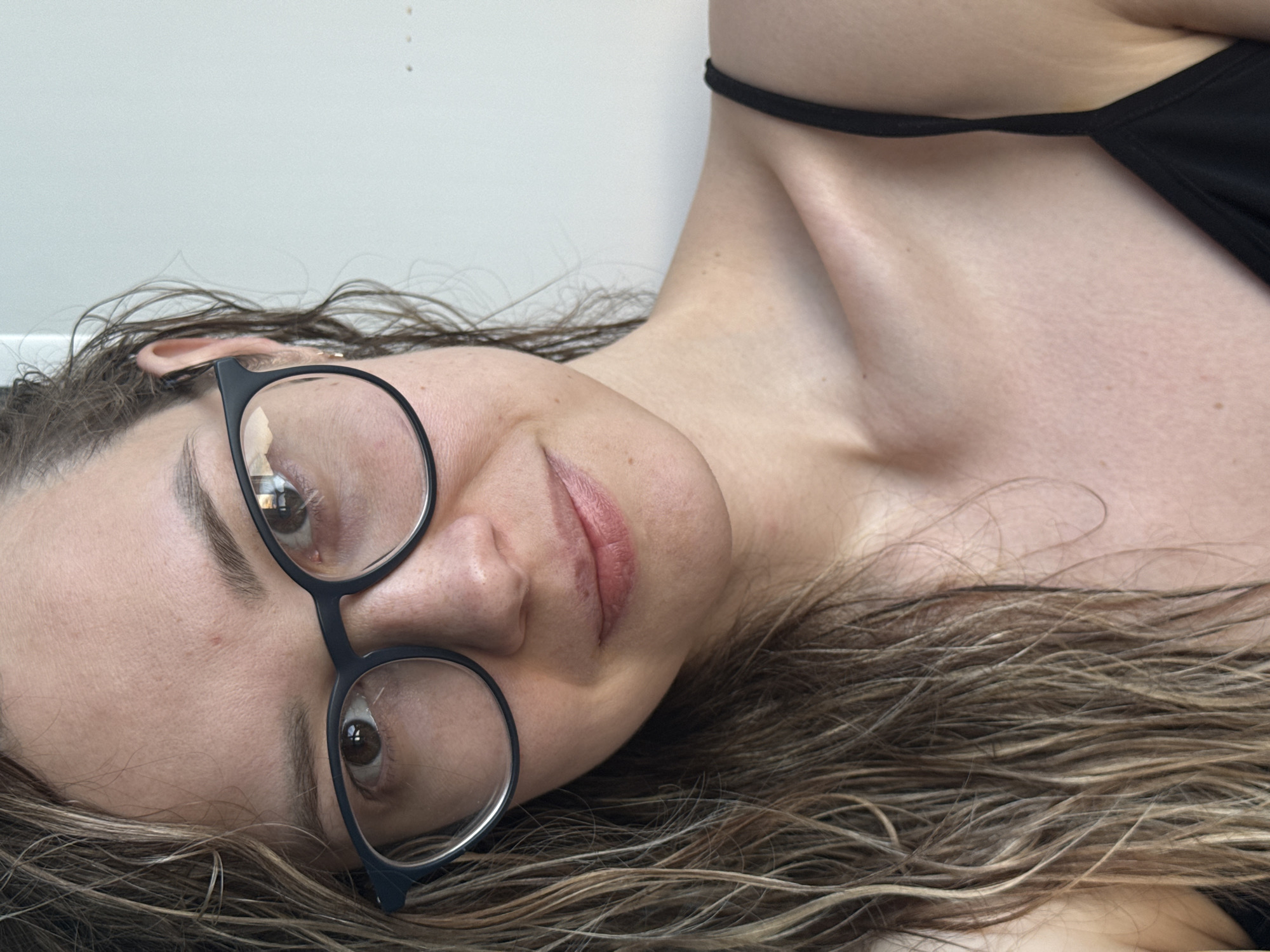 A person lies on their side. They have light skin and are wearing glasses with black frames. The person's hair is light brown and wet. The person is wearing a black top. The background is a plain white wall. There is no information to place the location in the city of Chaikovsky, Russia.