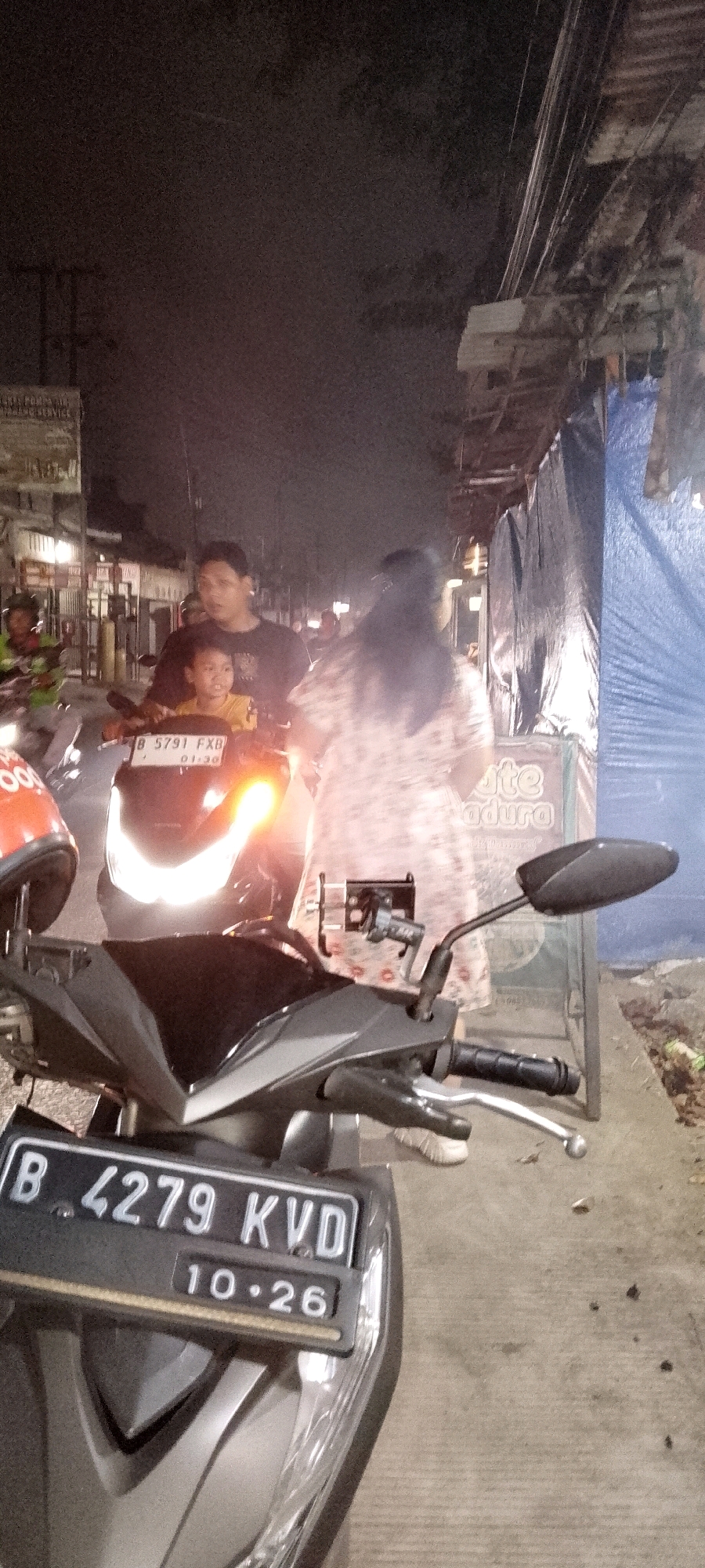 A low-light, full shot captures a family on a scooter at night on a street. The scooter, a dark grey Honda, has a license plate reading "B 4279 KVD" and a smaller plate with "10.26." The driver is a man with dark hair, wearing a black t-shirt. He is looking forward. In front of him, a young boy with short dark hair, wearing a yellow t-shirt, is seated. He is looking to his left. Partially visible to the right of the scooter is a woman with long dark hair, wearing a patterned dress. She is looking away from the camera. A blue tarp is visible on the right side of the frame, behind the woman, and a sign that says "Lite Madura" is on the tarp. The background shows a street with some buildings and electrical wires overhead. The street surface appears to be asphalt or concrete. The overall scene suggests a typical street in Satriamekar, Indonesia, at night, with people commuting on motorcycles.