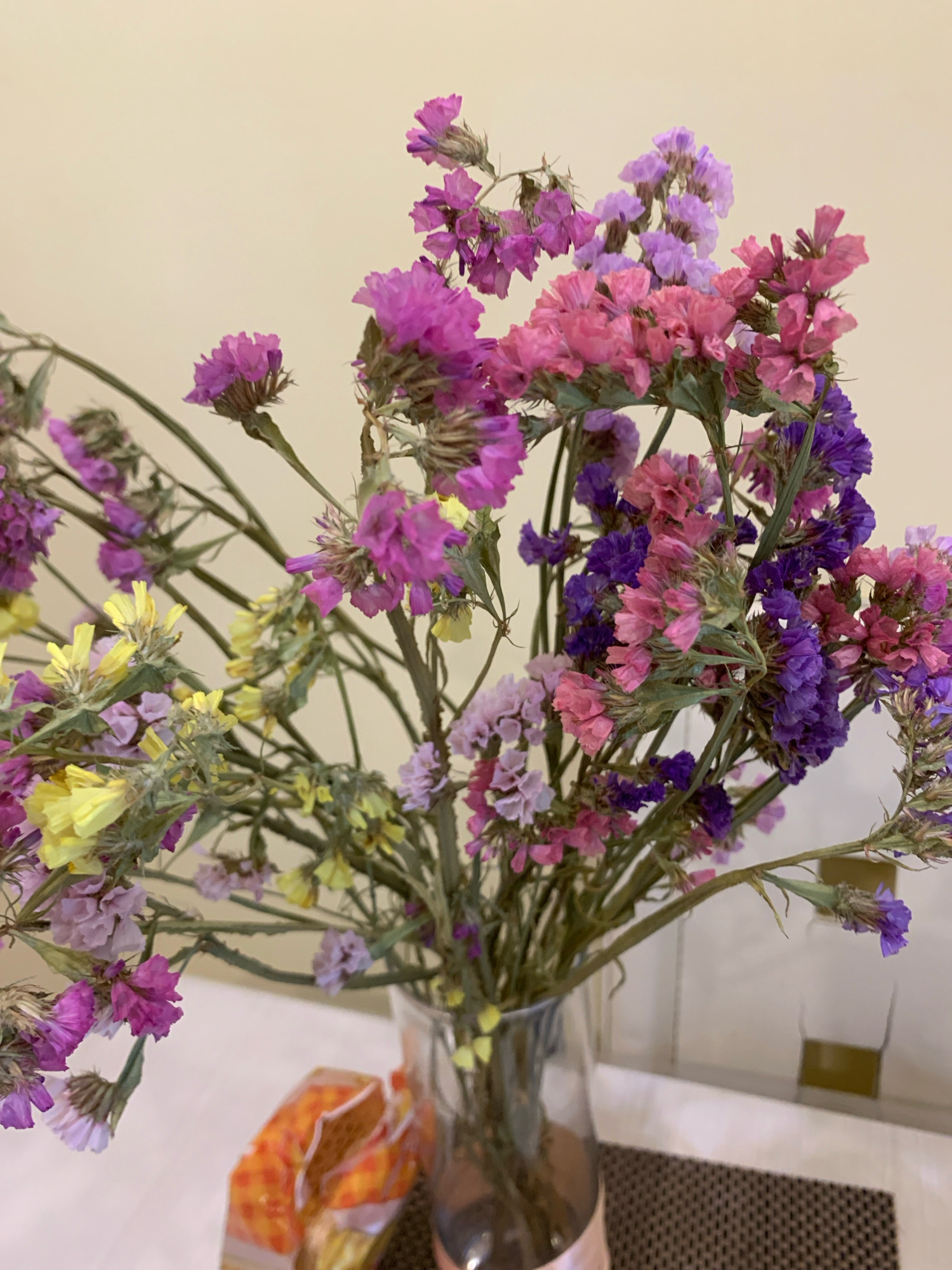 A bouquet of dried statice flowers in various shades of pink, purple, and yellow is displayed in a clear glass vase. The vase is positioned on a light-colored surface, possibly a table, with a textured placemat underneath. In the foreground, to the left of the vase, is a partially visible orange and yellow package, possibly containing snacks or food items. The background consists of a plain, light-colored wall. The scene appears to be indoors, in a domestic setting, likely a room in a house or apartment. There are no people or animals present in the image. The lighting suggests it could be daytime, with artificial lighting potentially augmenting the natural light. The overall impression is a still life composition focusing on the dried flowers. The specific location in Minsk, Belarus cannot be determined from the visual information provided in the image.
