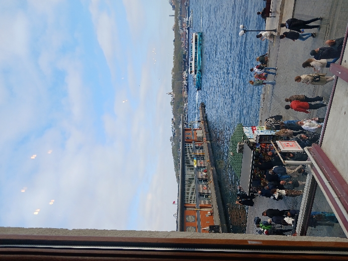 A view from a window overlooking the Golden Horn in Istanbul, Türkiye (41.022, 28.976). The image shows a ferry in the water and a number of people walking on the promenade in front of a building with a red roof and large windows. The sky is blue with a few white clouds. The image is taken from a building, likely a cafe, where the viewer can see people sitting at tables and the view through the window. Several people are looking out towards the water, which is calm and reflecting the blue sky. A few small boats are docked in the water behind the ferry. The environment appears to be a busy, bustling urban area, with the promenade, ferry, and cafe suggesting a popular tourist destination. The building in the background is likely a ferry terminal, with a red flag flying from its roof.