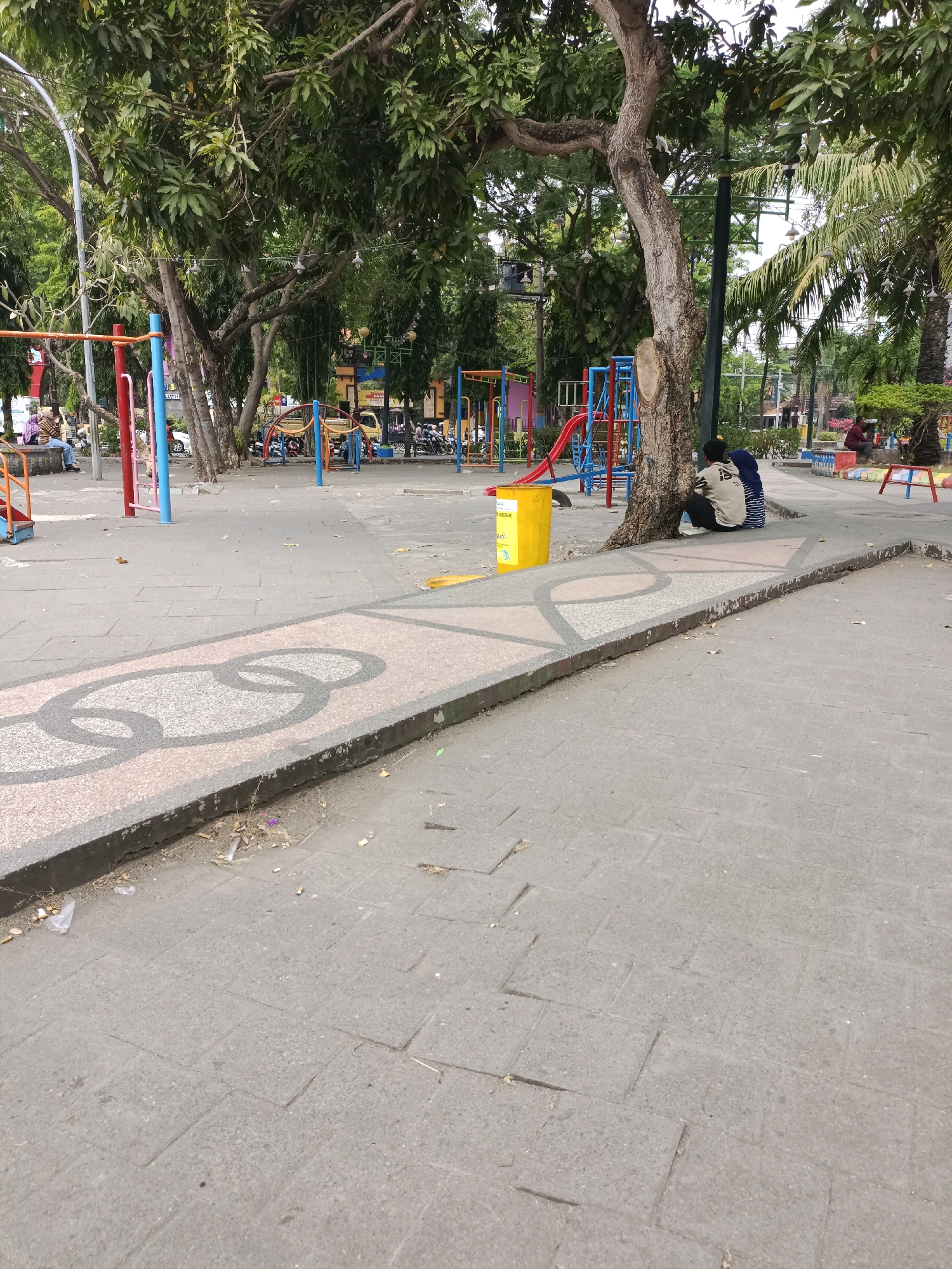 The image depicts a public park in Lamongan, Indonesia, likely a children's play area. The ground is paved with grey rectangular stones, with decorative sections featuring an interlocking ring pattern in beige and grey. Lush green trees dominate the background, with some palm trees also visible.
In the mid-ground, a bright yellow trash bin stands near the base of a large tree. Two individuals are seated on the ground, leaning against the tree trunk. The person on the left wears a light-colored long-sleeved shirt and dark pants, while the person on the right wears a blue headscarf and a dark garment. They appear to be interacting or resting together.
Further into the park, a colorful playground structure with red and blue elements and a slide is visible. Several other playground items, including swings and climbing frames, are scattered throughout the area. Various vehicles, including motorcycles and what appears to be a yellow car or van, are parked or moving in the background, suggesting the park is accessible to the public and situated near a road or parking area. Some people are visible sitting on a low wall on the left side of the image. The overall scene conveys a sense of a moderately busy public space on a day with natural lighting.