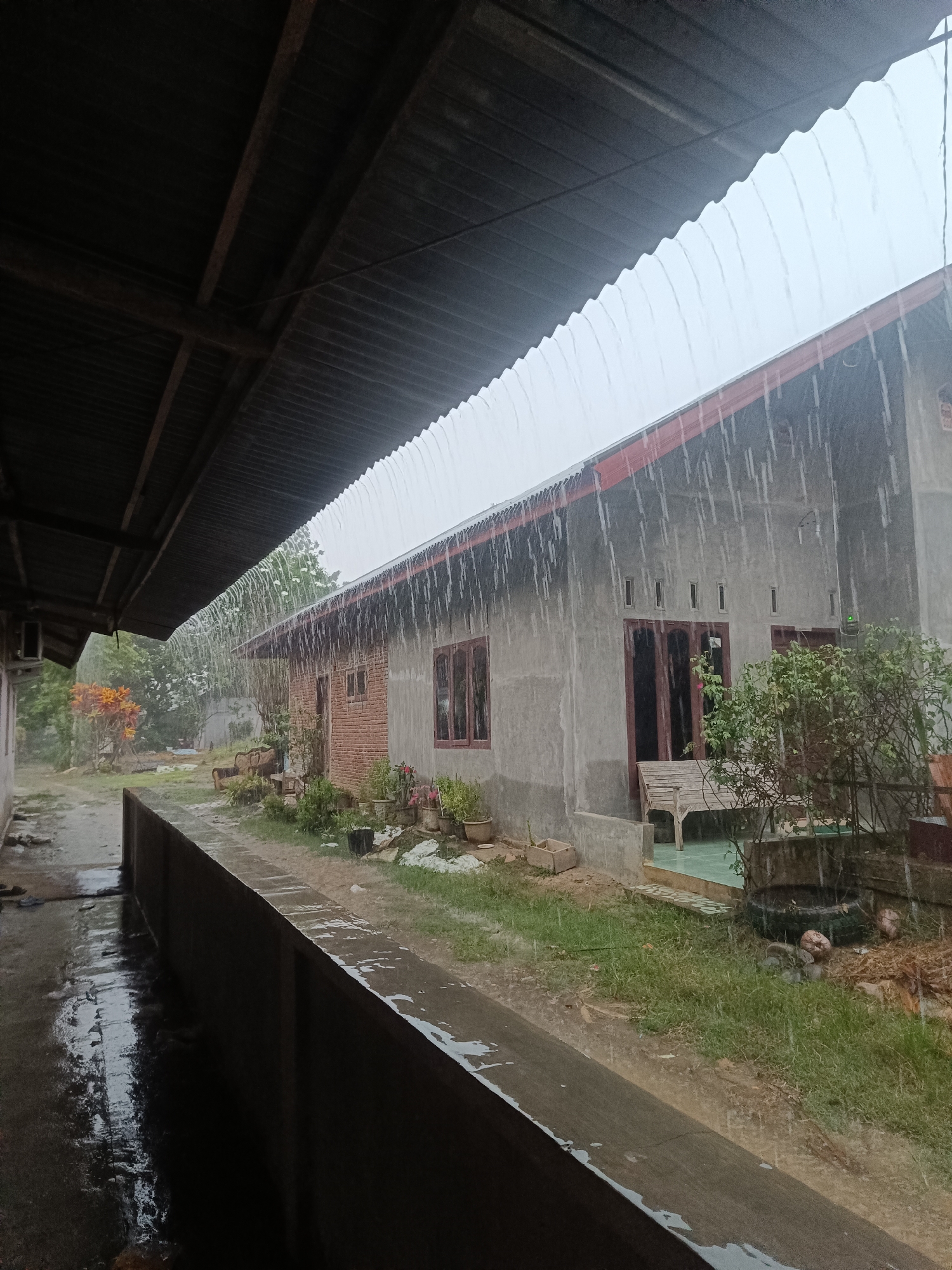 The image shows a heavy downpour in Aluebuket, Indonesia. Two buildings are visible, one with brick and concrete construction, and the other with a corrugated metal roof extending over a walkway. Rainwater streams down heavily from the eaves of both structures. Vegetation is present, including small trees and potted plants near the buildings. Water accumulates on the ground and walkway surfaces. No people are visible.