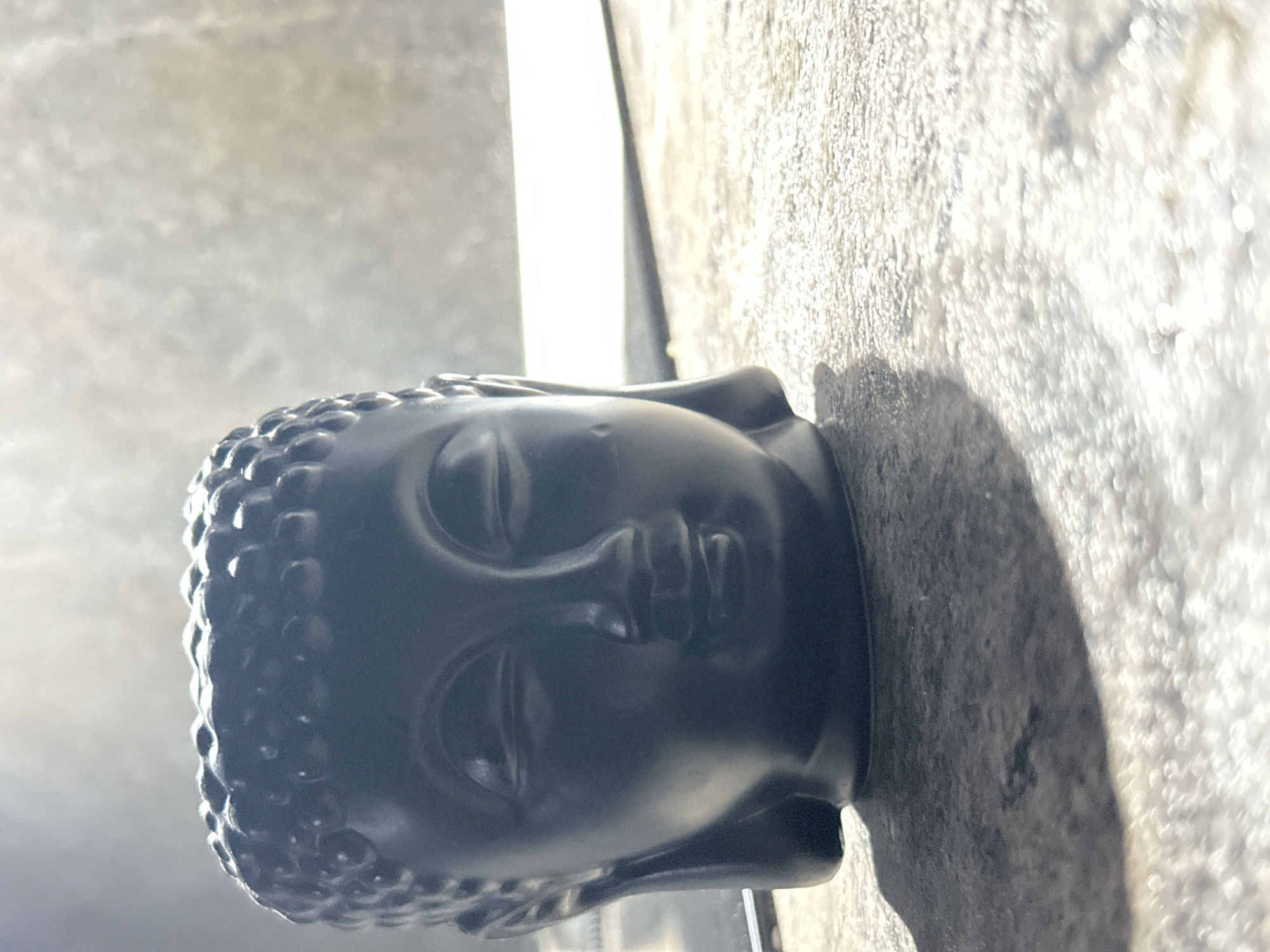 A close-up, eye-level shot shows a dark gray Buddha head statue resting on a rough, light gray surface. The statue is the primary focus, with detailed features of the face and the textured "hair" characteristic of Buddha representations. A distinct shadow is cast to the right of the statue on the surface.
The surface appears to be a stone or concrete ledge. A blurred, light background suggests an outdoor setting, possibly a balcony or patio. No people or other objects are visible in the immediate vicinity. The location in Krasnodar, Russia, is not discernible from the visual information.