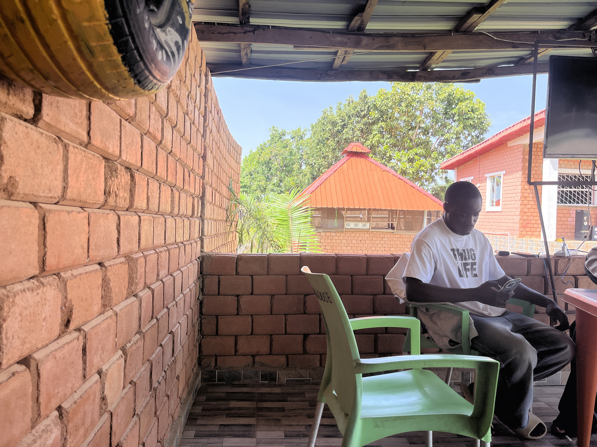A man in a white t-shirt with "THUG LIFE" printed on it is seated in a green plastic chair, looking at his mobile phone. He is wearing dark pants and light-colored slides. The scene is an outdoor, covered area with a brick wall on the left and a corrugated metal roof supported by wooden beams. In the background, there are trees, a building with an orange roof, and another red-brick building with white trim and a window. The man is positioned in what appears to be a seating area or patio. The environment suggests a warm climate, consistent with Jos, Nigeria. The overall lighting is bright, indicating daylight.