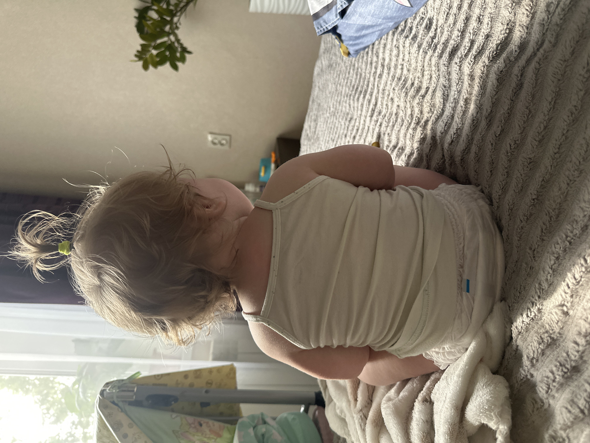 A toddler with blonde hair tied into a high ponytail sits on a bed facing away from the camera. The toddler is wearing a white tank top and a diaper. The bed is covered with a textured grey blanket and a cream-colored fluffy blanket is draped over the left side. In the background, a window with sheer curtains allows natural light to enter the room. To the left of the window, a baby crib with a patterned quilt is visible. A light switch is mounted on the wall behind the toddler. A potted plant with green leaves sits on a nightstand to the right. The overall setting suggests a domestic environment, likely a bedroom, in the city of Sayanogorsk, Russia.